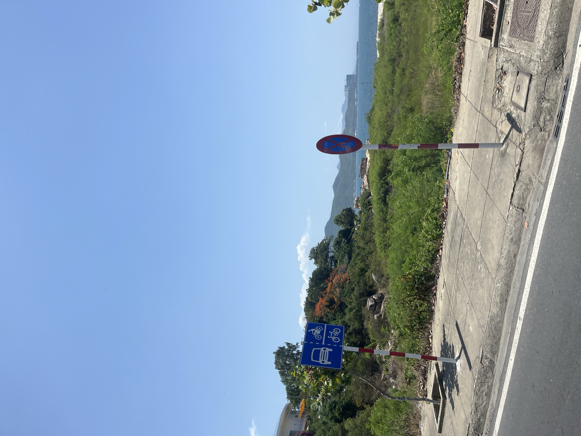 The image shows a street scene in Nha Trang, Vietnam, with a clear blue sky overhead. In the foreground, a concrete sidewalk runs alongside a road. Two traffic signs are visible. The first sign, on the left, is blue with white symbols depicting a car, a motorcycle, and a bicycle, indicating a route for these vehicles. It is mounted on a pole with red and white stripes. The second sign, on the right, is circular with a red border and a blue center, featuring an "X" symbol in red, which typically signifies a no stopping or no parking zone.
Behind the sidewalk, lush green vegetation covers a sloping hillside, dotted with rocks. Several trees are interspersed within the greenery, some with vibrant orange flowers. In the distance, the sparkling blue sea stretches to the horizon, meeting a hazy range of mountains under the bright sky. A few buildings are visible along the coastline, including a tall structure under construction. The overall environment suggests a coastal road with a mix of urban and natural elements.