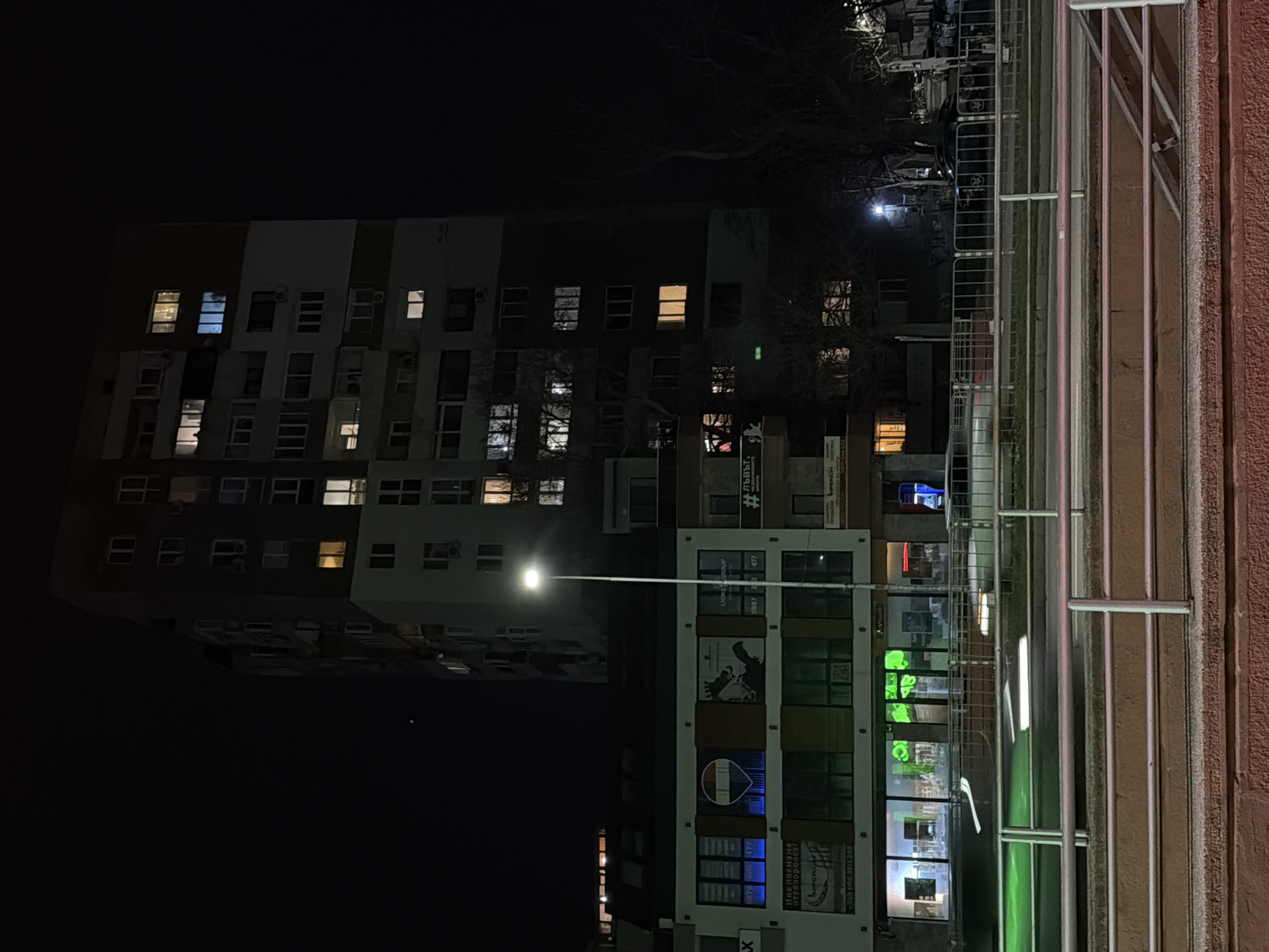 The image is a night scene in Varna, Bulgaria, featuring a tall apartment building on the left with numerous illuminated windows. Adjacent to it is a commercial building with storefronts, including those displaying advertisements and green signage. A street lamp casts a glow between the buildings. In the foreground, a metal railing runs along a reddish-brown wall.