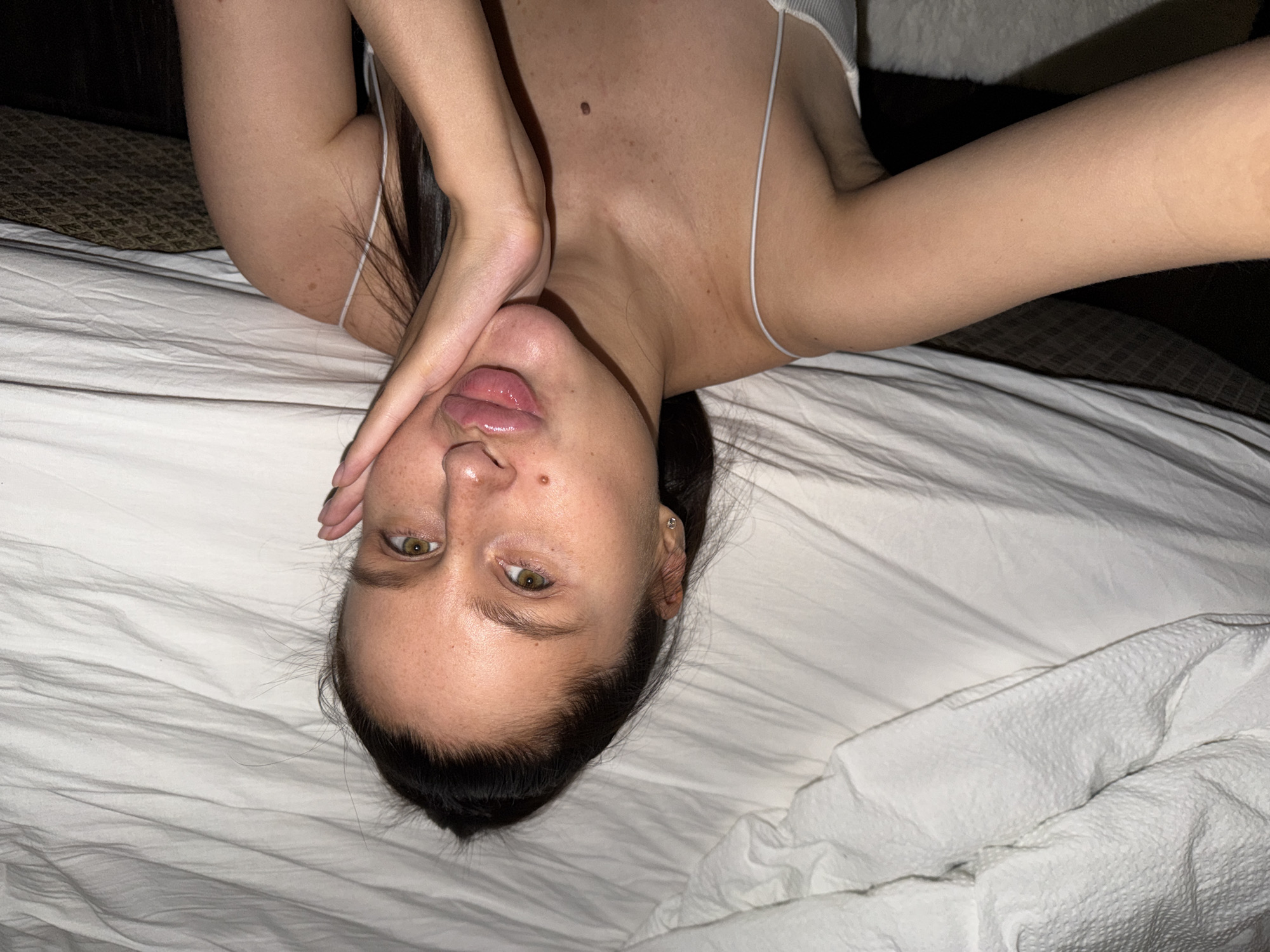 An overhead, close-up photograph captures a young adult female lying on a bed, positioned upside down in the frame. The setting appears to be an indoor space, likely a bedroom in Khimki, Russia, characterized by crumpled white bed sheets. The lighting is soft and somewhat dim, suggesting evening or nighttime.
The subject has fair skin, with visible freckles across her face and chest. Her dark hair is pulled back, accentuating her green or hazel eyes, which are looking directly at the camera. Her lips are slightly parted and appear glossy. One hand is raised, with fingers curled around her chin and mouth, adding a pensive or playful element to her pose. She wears a light-colored top with a thin white spaghetti strap visible over her right shoulder. A small stud earring can be seen in her right ear.
Her arms are outstretched to the sides, with one arm extending towards the top left of the frame. A dark, patterned object, possibly a pillow or headboard, is partially visible behind her upper left shoulder. The image focuses closely on her face and upper torso, conveying a sense of intimacy. No visible text is present in the image.