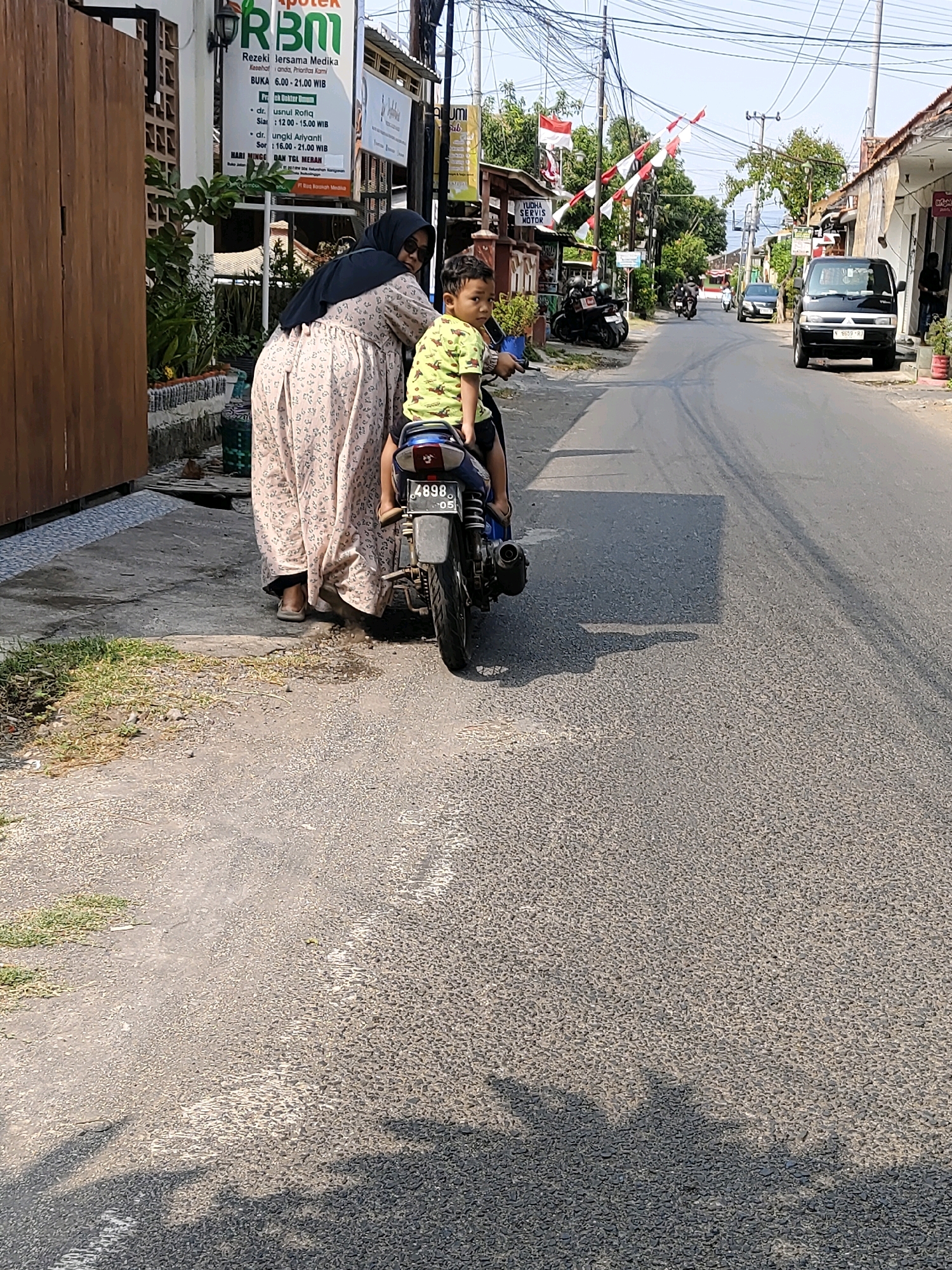 The image shows a street scene in Probolinggo, Indonesia. In the foreground, a woman wearing a headscarf and a long floral dress is standing next to a motorcycle. A young boy is seated on the back of the motorcycle, looking towards the camera. The woman appears to be holding onto the motorcycle. The street is paved and has some markings. On the left side of the street, there is a wooden fence and a building with a sign that reads "APOTEK RBMI". Further down the street, there are other buildings, trees, and more motorcycles. Indonesian flags are strung across the street in the middle ground. A white van is parked on the right side of the street. The scene appears to be during daylight hours.