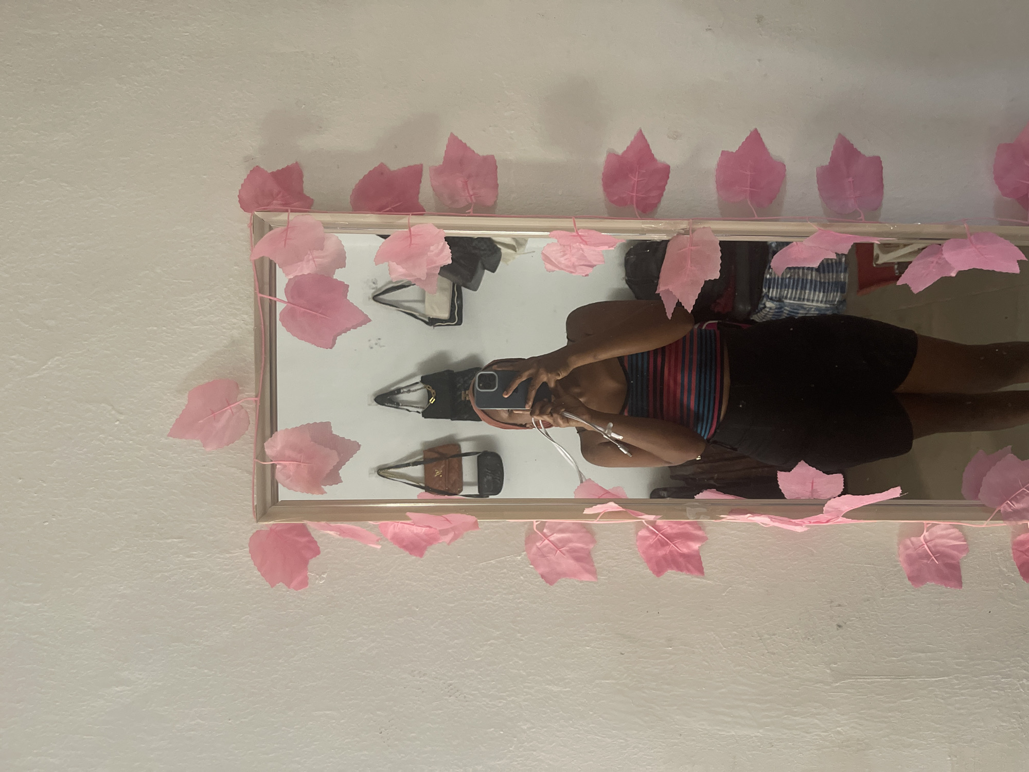 The image depicts a young woman standing in front of a mirror, taking a selfie. She is wearing a striped top and black shorts, with a pink headband. The mirror is adorned with a string of pink artificial leaves. In the reflection of the mirror, several handbags are visible hanging on the wall. The background appears to be a simple white wall. The scene is indoors, likely in a bedroom or dressing area. The image does not contain information that would help determine the specific location within Rafin Par, Nigeria.