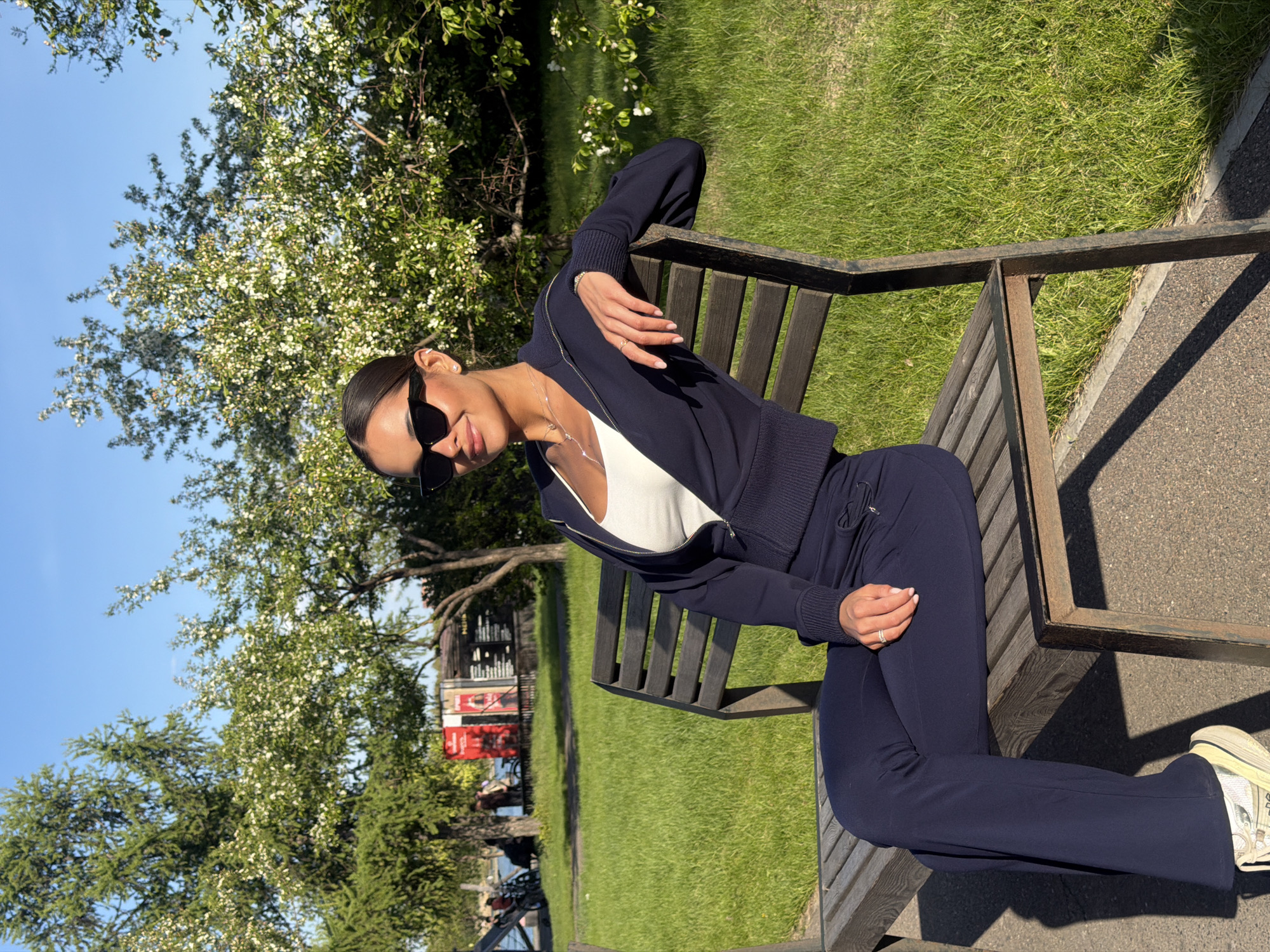 A woman wearing a navy blue tracksuit and sunglasses sits on a wooden bench in a park. The park appears to be located in Krasnoyarsk, Russia, judging by the street signs in the background. The woman is wearing a white top underneath her jacket. The background includes green grass, trees with white flowers, and a paved walkway. The day appears sunny.