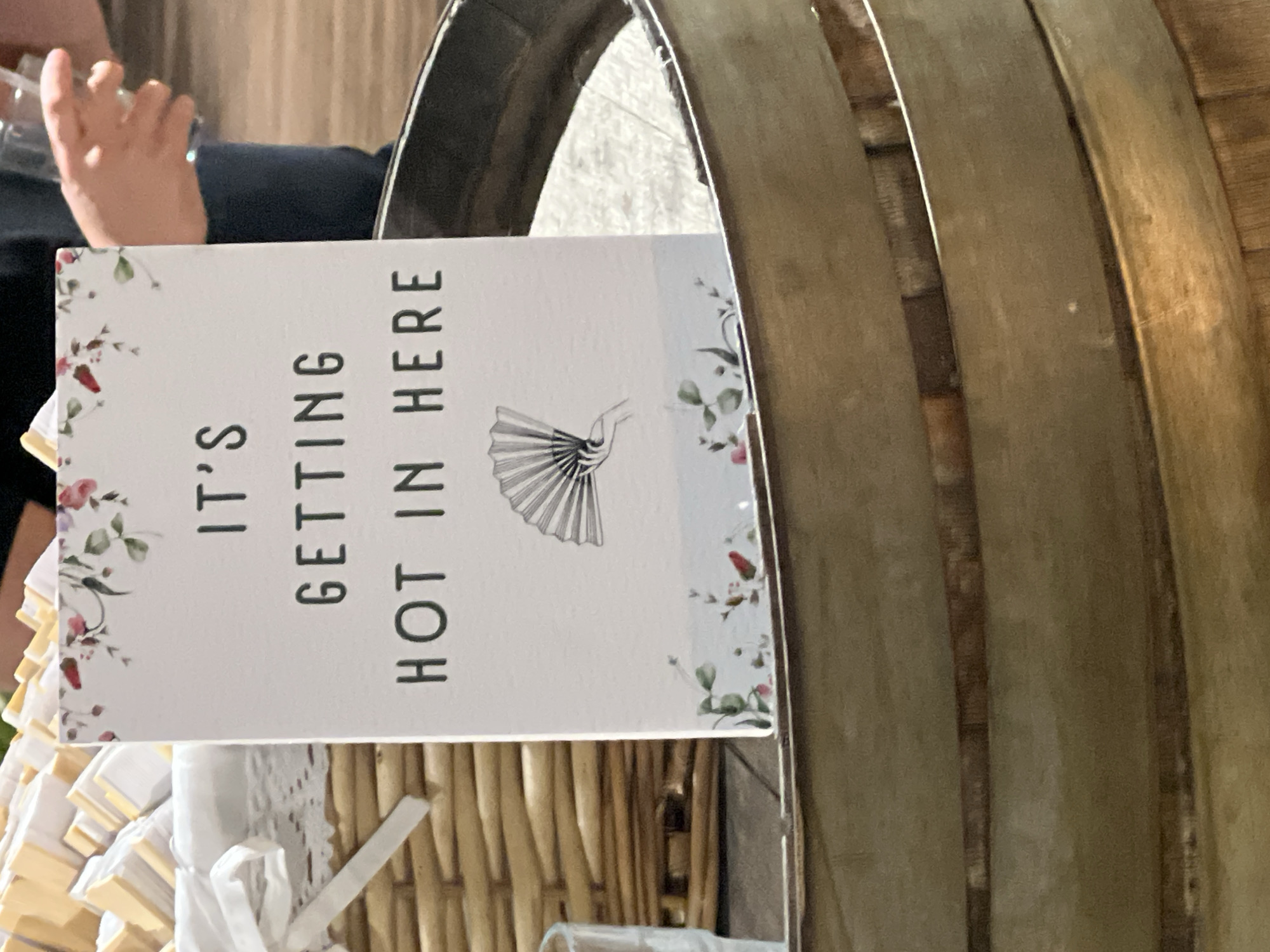 This is a close-up, eye-level, indoor shot of a sign that reads "IT'S GETTING HOT IN HERE" in dark lettering. The sign is white with a floral border at the top and a faded illustration of a hand holding a fan at the bottom, all set against a pale blue background with faint floral accents. The sign is propped inside a dark, circular wooden barrel. To the left of the sign is a woven basket filled with small, individually wrapped items, possibly favors or treats, tied with white ribbons. The background shows a person's hands holding a clear plastic cup. The setting appears to be a venue in Beit Hanan, Israel, possibly a wedding or event. The lighting is natural and somewhat diffused.