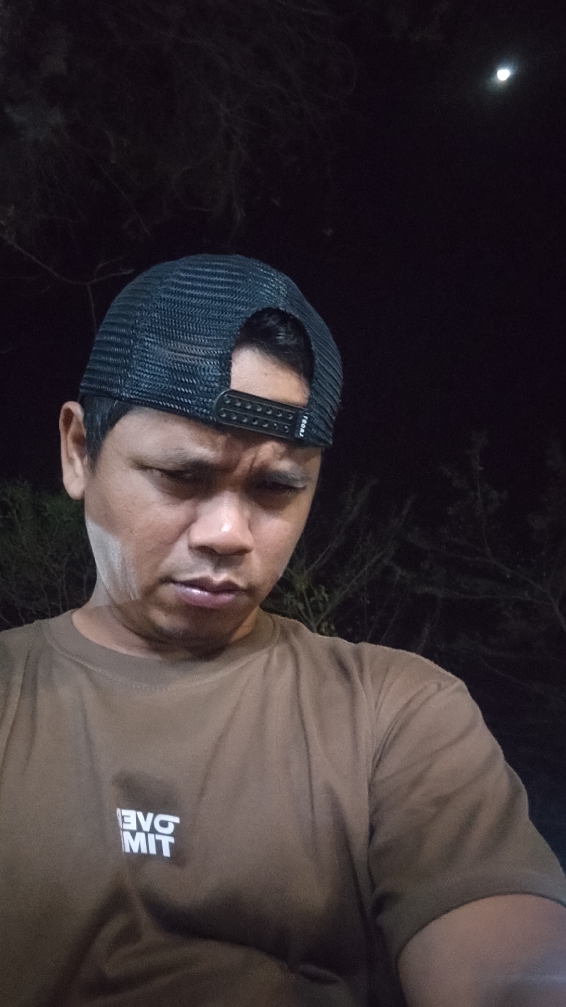The image is a close-up, waist-up portrait of a man wearing a black trucker hat and a brown t-shirt. He is looking down with a pensive expression, and there is a white mark on his left cheek. The background is dark, with some blurry foliage and a faint moon visible in the upper right corner. The man is in Karanglangu, Indonesia, but no specific location within the city is provided.