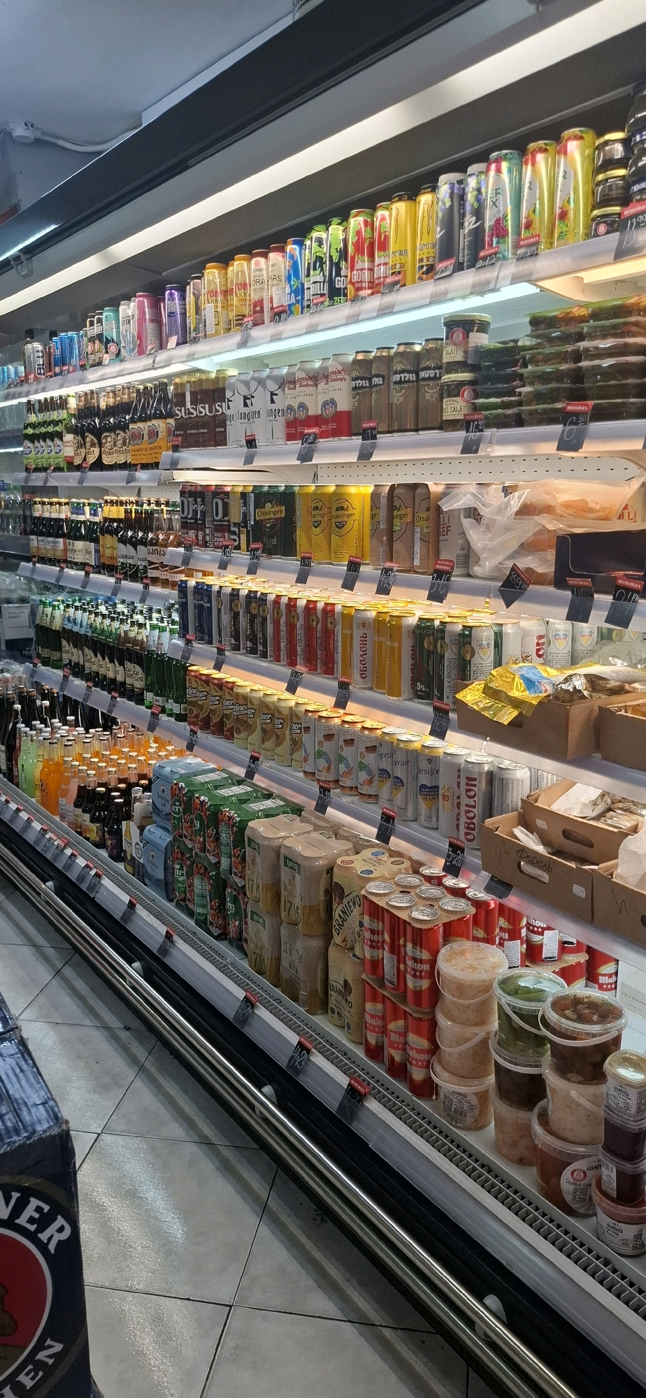 The image displays a well-lit aisle of a supermarket in Yerocham, Israel, specifically showcasing a refrigerated display case filled with various beverages and food items. The shelves are neatly organized with rows of canned drinks, including beer, soda, and energy drinks, identifiable by their colorful labels and packaging. Several brands are visible, such as "Oettinger," "Mahou," and "Monster Energy." Among the beverages, there are also various food items packaged in plastic containers, including what appear to be pickled vegetables and spreads. The overall environment suggests a modern retail setting, with clean floors and efficient shelving systems designed for product visibility and accessibility. The lighting is bright and even, illuminating the products clearly. No people are visible in the image.