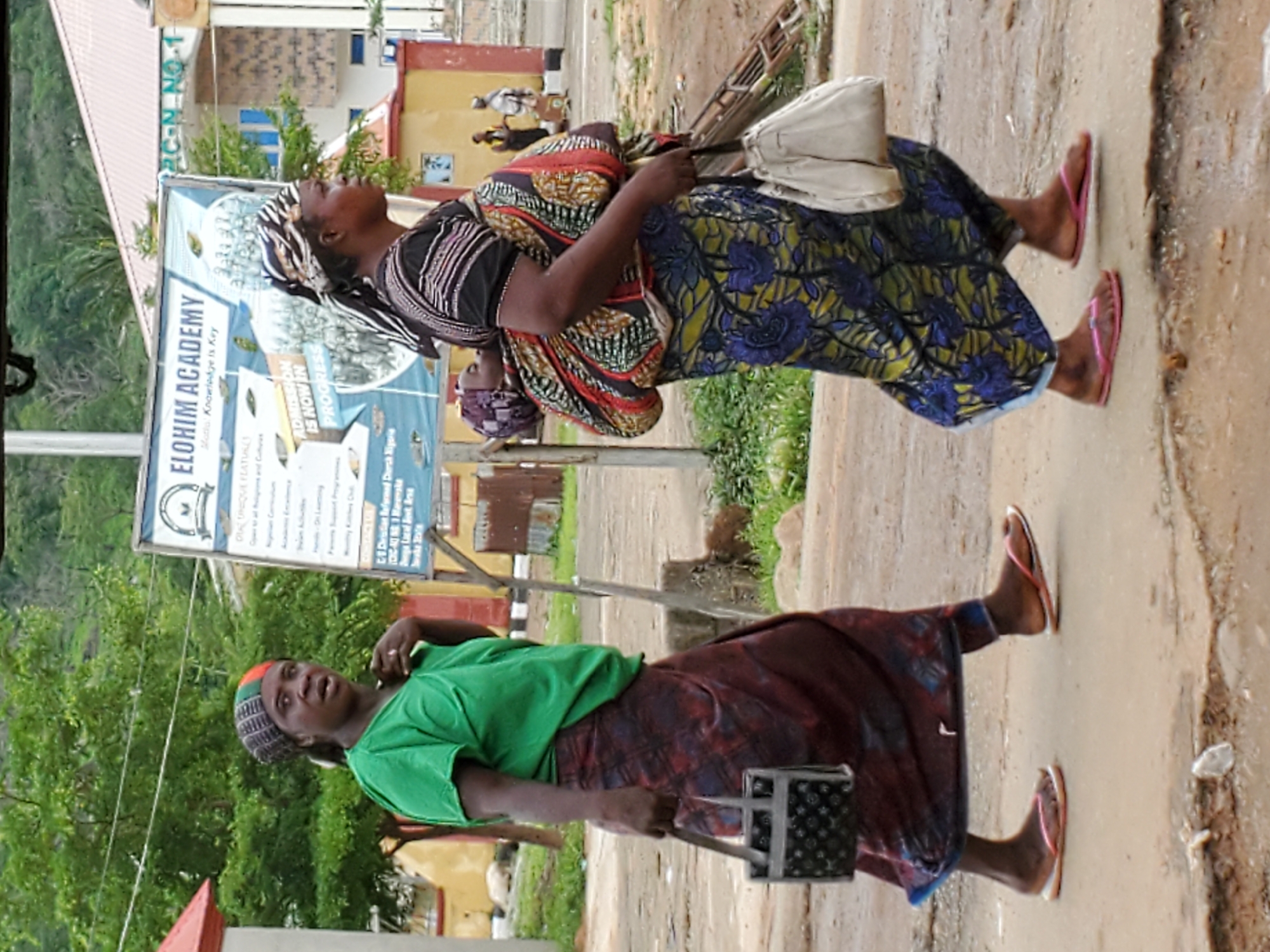 The image depicts an outdoor scene in Mararaba, Nigeria, showing two women walking along a dirt or unpaved path during the daytime.
In the foreground on the left, a woman wears a green t-shirt, a multi-colored knitted cap, and a dark patterned skirt, with pink flip-flops on her feet. She holds a small, dark, textured container with a handle and looks towards the viewer with a neutral or attentive expression.
On the right, another woman is dressed in a patterned headscarf, a striped top, and a long, dark blue skirt with floral patterns, also wearing pink flip-flops. She carries a light-colored handbag over her shoulder and another patterned fabric bag draped over her arm, her head tilted back as if looking skyward.
The background features lush green foliage, including trees and bushes, suggesting a natural environment. Several buildings with painted walls and corrugated roofs are visible. A large billboard prominently displays text, identified by OCR as "ELOHIM ACADEMY" with the motto "Knowledge is Key" and "ADMISSION IS NOW IN PROGRESS." Further details on the sign mention "Christian Reformed Church Nigeria" and snippets like "1 Maret" and "Lagal Best Arsa." The letters "RC-N NO-1" are visible on a nearby building. The ground appears somewhat damp or muddy in places, but the overall lighting suggests a bright, possibly sunny, day. The image captures a moment of daily activity in a local community.