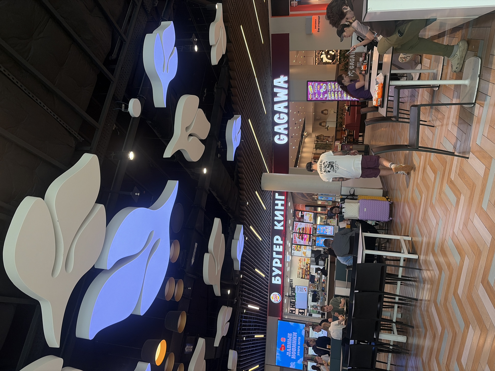 The image depicts a food court area, likely within a shopping mall, with a Burger King and a Gagava restaurant visible. The environment is brightly lit with modern, stylized ceiling decorations that resemble large white leaves, some of which are illuminated with blue light. The floor is tiled in a herringbone pattern of wood-toned and light grey tiles.
In the foreground, several tables with chairs are arranged. A man wearing a white t-shirt with graphics and purple shorts walks away from the camera. A purple suitcase and a yellow suitcase are visible near a table on the right.
Further back, patrons are seated at tables in both Burger King and Gagava. In the Burger King section, a large screen displays "Love is..." and "ЛАВНЫЕ НОВИНКИ этого лета" (Love is... and The Hottest Newcomers of This Summer). A menu board with images of food items is displayed above the counter.
In the Gagava section, a vertical digital menu board shows food items and prices, with "PANCHI 430" being partially legible.
The overall scene suggests a busy food court, likely in Saint Petersburg, Russia, given the Russian text on the Burger King signage and the digital menu. The atmosphere appears casual and functional, typical of a public dining space.