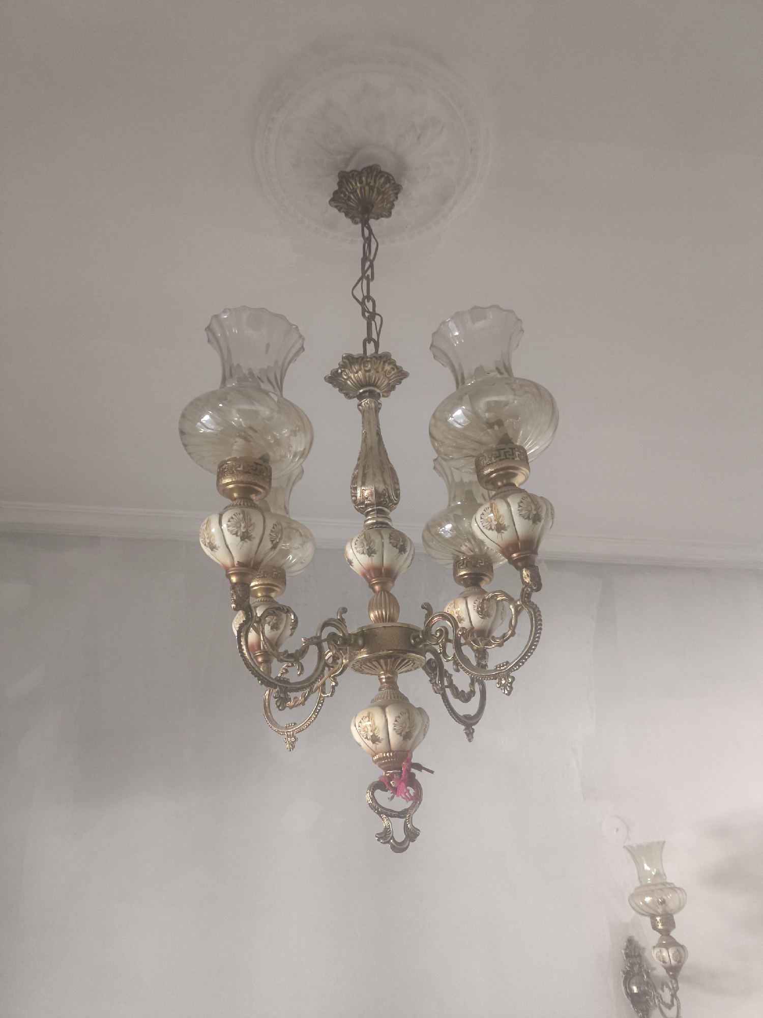 A vintage, ornate chandelier hangs from a ceiling with decorative molding. The chandelier is made of brass with intricate scrolling arms supporting six lamps. Each lamp has a clear, fluted glass shade and a decorative, bulbous ceramic base with floral painted details. The chandelier is illuminated by light bulbs. To the right and lower, a wall sconce with a similar design is visible. The walls are painted a light color. The scene is indoors, likely in a home in Petroupoli, Greece.