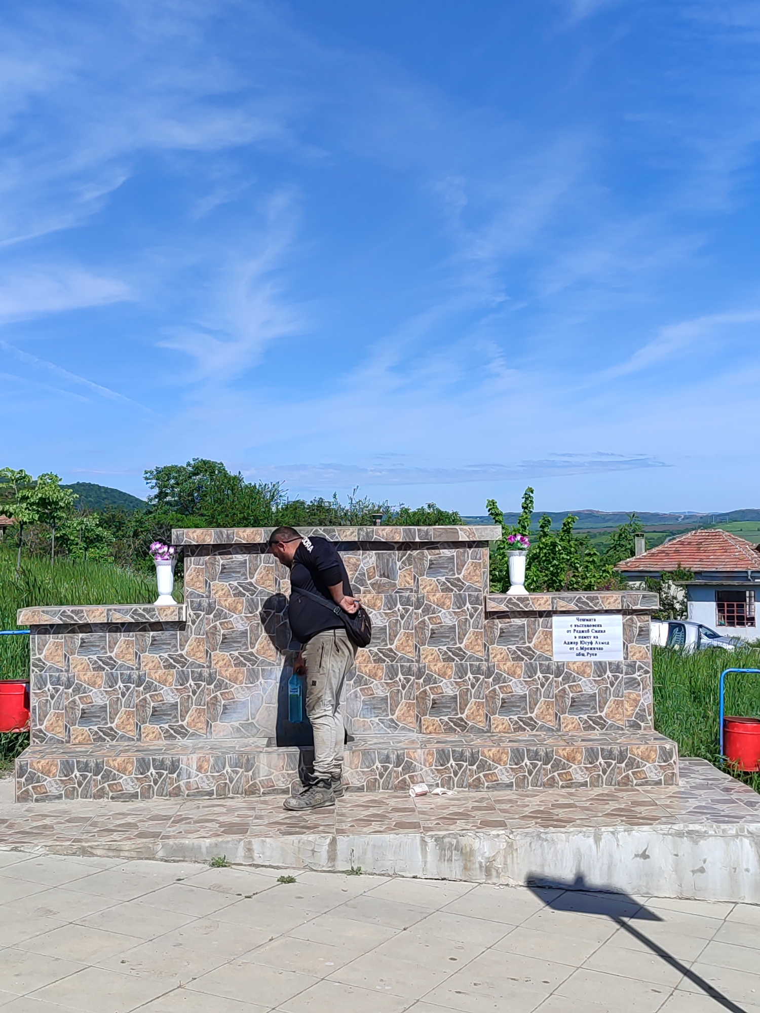 A man in a black shirt and tan pants is standing at a stone fountain on a clear day. He is bent over, appearing to fill a blue water bottle. The fountain is tiled with a mosaic pattern and has white vases with flowers on either side. There is a sign on the fountain with text written in Bulgarian, as well as red buckets attached to the structure. In the background, there is a rural landscape with green hills and a blue sky with wispy clouds. The location is possibly near Medovo, Bulgaria.