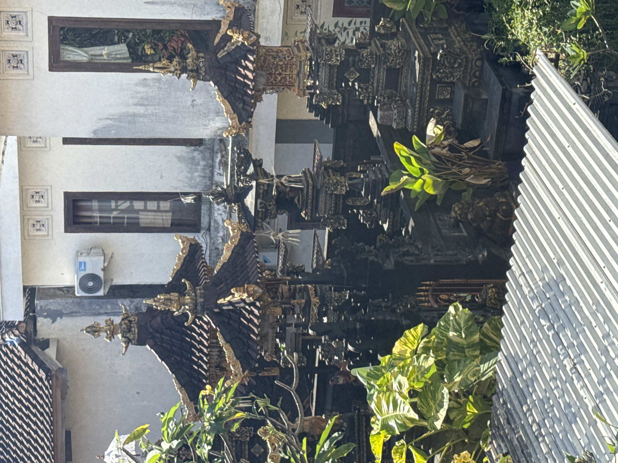 An eye-level, medium shot depicts a building with ornamental structures in Kuta, Indonesia. The building is off-white, and has two windows and an air conditioning unit, with light entering. The windows are rectangular, with a dark brown frame. The air conditioning unit is white with blue details. In front of the building, several black tiered ornamental structures are seen. These structures are decorated with gold accents and are partially obscured by plants and foliage. At the right side of the image, a section of corrugated metal fencing is visible, adding a modern contrast to the traditional architecture. The sky is bright, suggesting a sunny day.