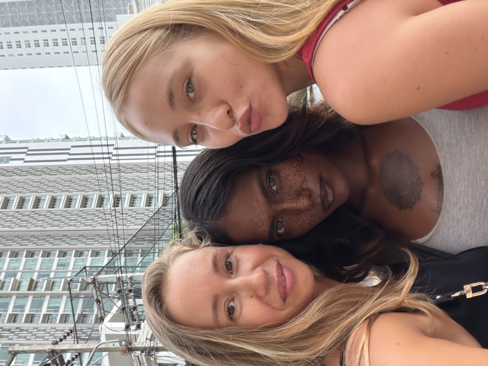 Three women are positioned closely together, likely taking a selfie. The woman at the top has blonde hair and is making a kissing face. The woman in the middle has dark hair and prominent freckles. The woman at the bottom also has blonde hair and is smiling. They appear to be outdoors in Pattaya City, Thailand, given the presence of a tall building and power lines in the background. The building has a light-colored facade with rows of windows. The woman in the middle has a tattoo on her upper chest/shoulder area.