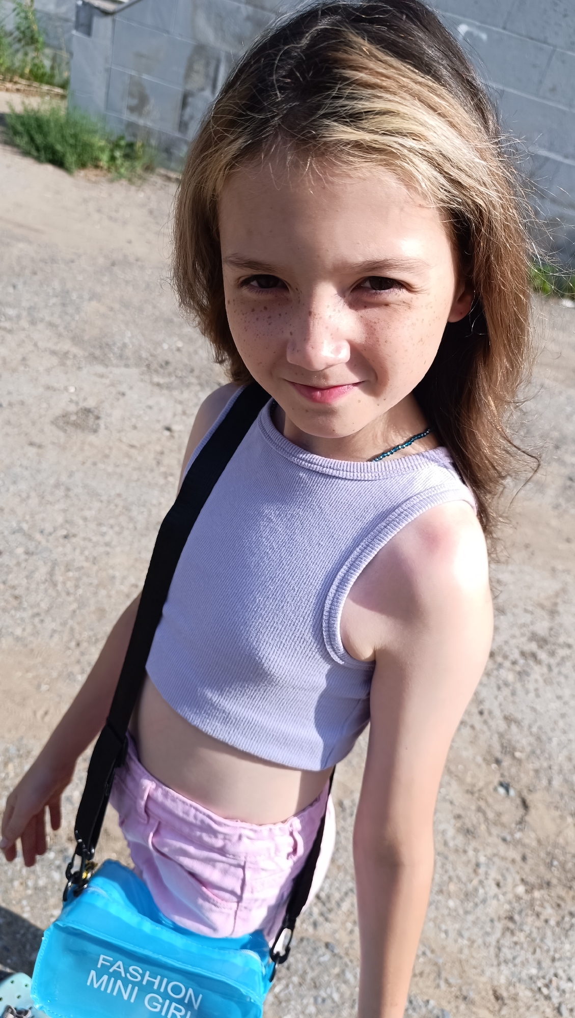 A young girl with freckles and long brown hair, styled with blonde highlights, looks directly at the camera with a slight smile. She is wearing a light purple, ribbed crop top and pink shorts. A black strap runs diagonally across her body, attached to a bright blue transparent bag with white text that reads "FASHION MINI GIRL". The girl has a turquoise beaded necklace around her neck. Her right shoulder appears slightly sunburned. She is standing outdoors on a gravelly surface, with a grey, textured wall and some green vegetation in the background. The lighting suggests it is daytime, likely sunny. The overall mood is casual and summery.