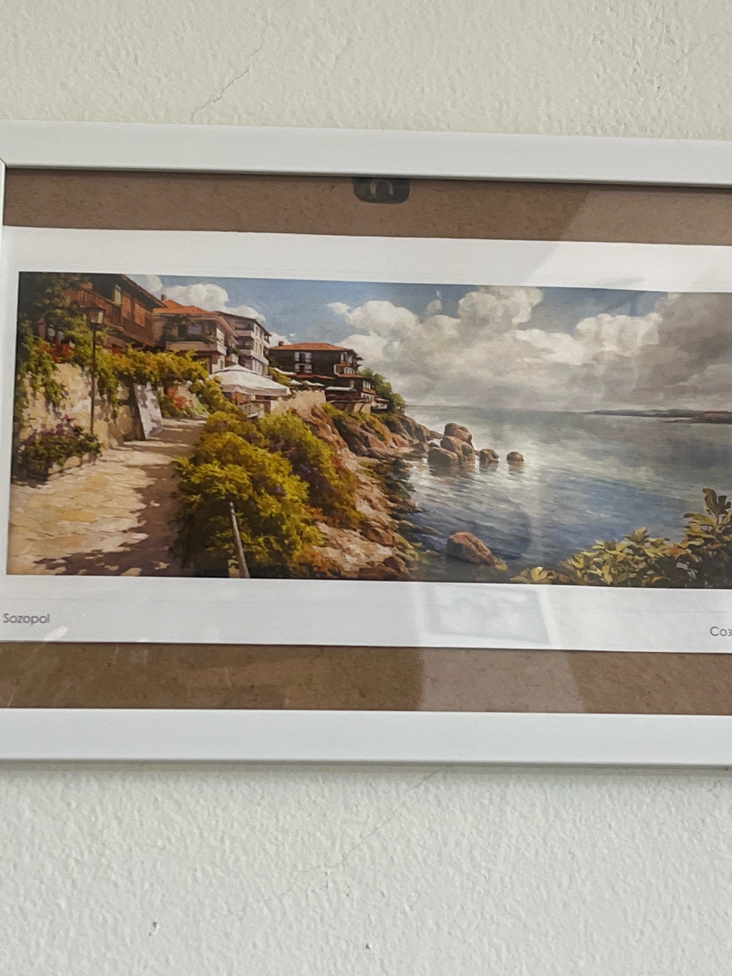 The image is a framed panoramic painting of a coastal town. The painting depicts a scenic view of buildings with terracotta roofs situated on a rocky coastline overlooking a body of water. Lush green vegetation covers parts of the rocky cliffs, and a paved pathway winds up towards the buildings. The water is calm with several rocks protruding from it, and the sky is filled with dramatic clouds. The bottom left corner of the painting has the word "Sozopol" printed in white, and the bottom right corner has "Cox" printed in white. Sozopol is a historic seaside town in Bulgaria. The painting is hung on a textured white wall.