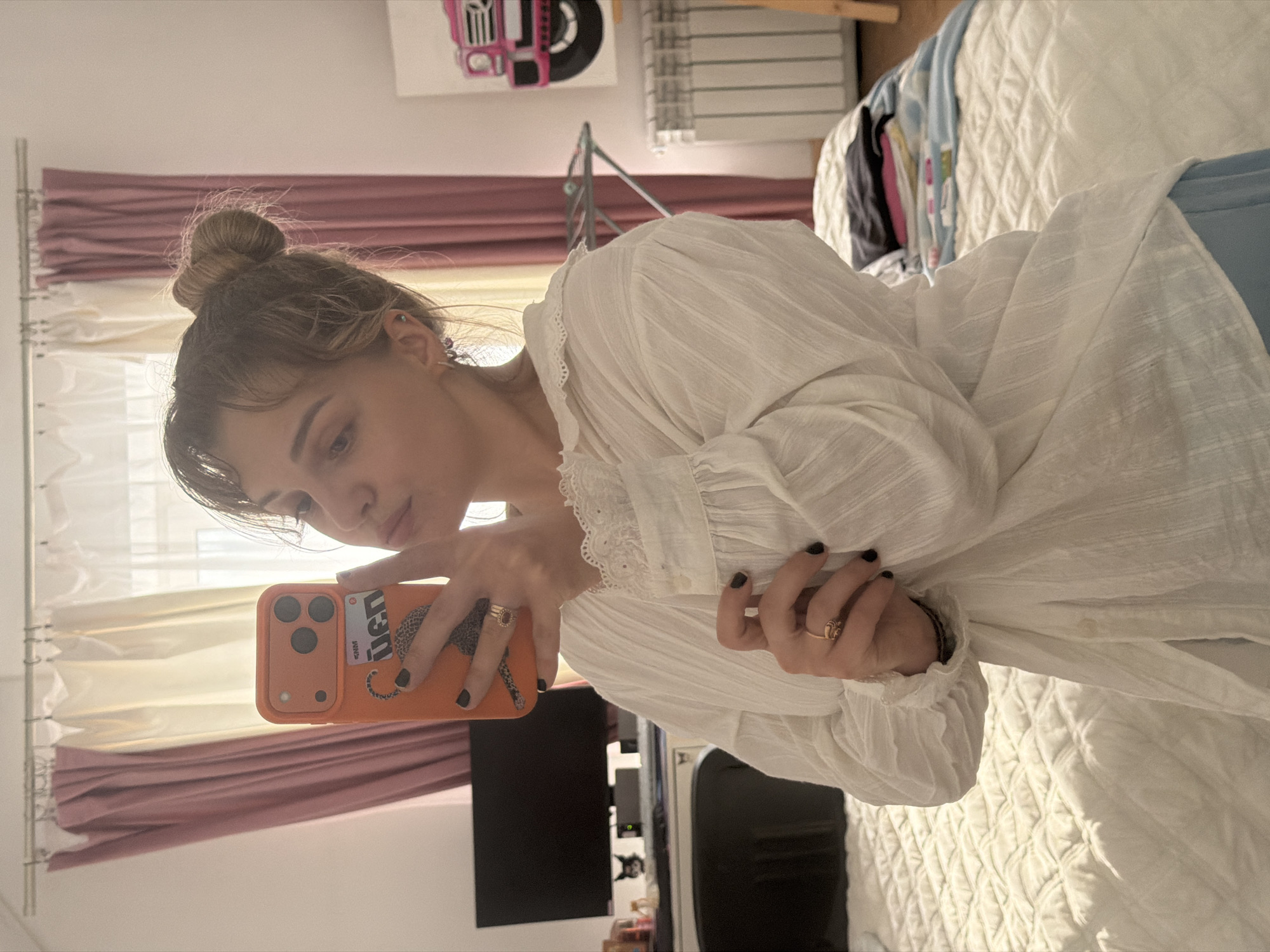A mirror selfie captures a young woman in an indoor setting, likely a bedroom, in Большая Шильна, Russia. She has light-colored hair styled in a high bun with some loose strands framing her face. Her expression is neutral, as she looks into the orange smartphone she holds up to take the photo. She wears a white, textured long-sleeved shirt featuring delicate lace details along the neckline or shoulders. Her fingernails are painted black or a very dark shade, and she wears several rings on her fingers, including a gold band with a reddish stone and another simpler gold ring. A small ear piercing with a gem is also visible.
The smartphone is encased in a distinctive orange cover adorned with a black cheetah or leopard print. Visible text on the case includes "DEAD" and "4MM" (or "eNM"). The phone itself appears to have a triple-lens camera system.
The background suggests a well-lit room, indicative of daytime. A window draped with white sheer curtains and an underlying layer of pink or mauve fabric is visible. Below the window, a white radiator is present. Behind the woman, a bed covered with a white, quilted comforter is partially visible, along with some folded clothes in blue, black, and pink hues. On the wall above the bed, a piece of artwork depicts a stylized pink and black front view of a vehicle, possibly a truck. A dark rectangular object, likely a television or computer monitor, is reflected in the lower part of the mirror.
