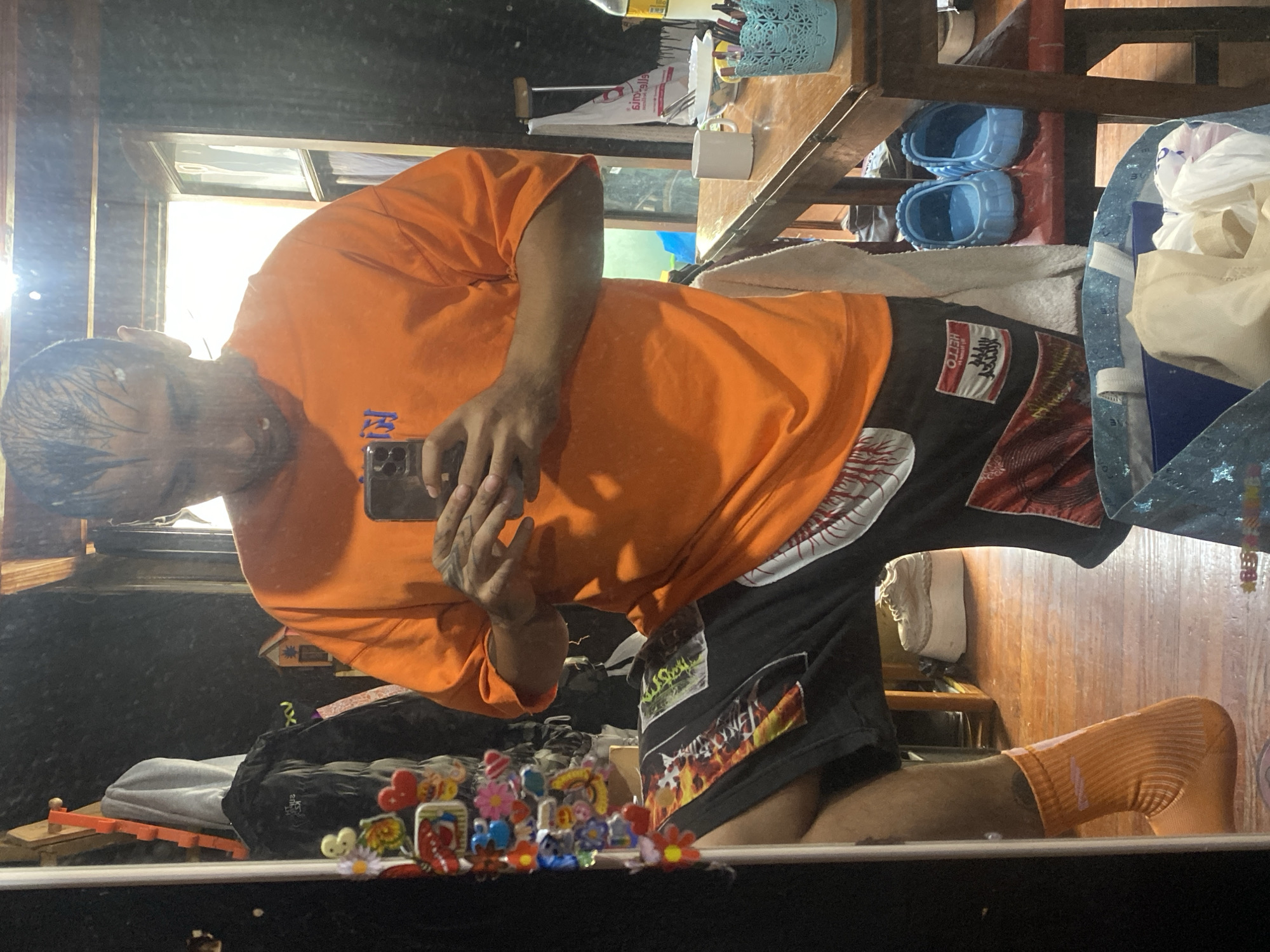 A man takes a photo of himself in a mirror. He is wearing an orange T-shirt and black shorts. His hair appears wet. The reflection in the mirror shows items on a shelf and a wooden floor. The location cannot be identified as Buenos Aires, Argentina, from the provided image.