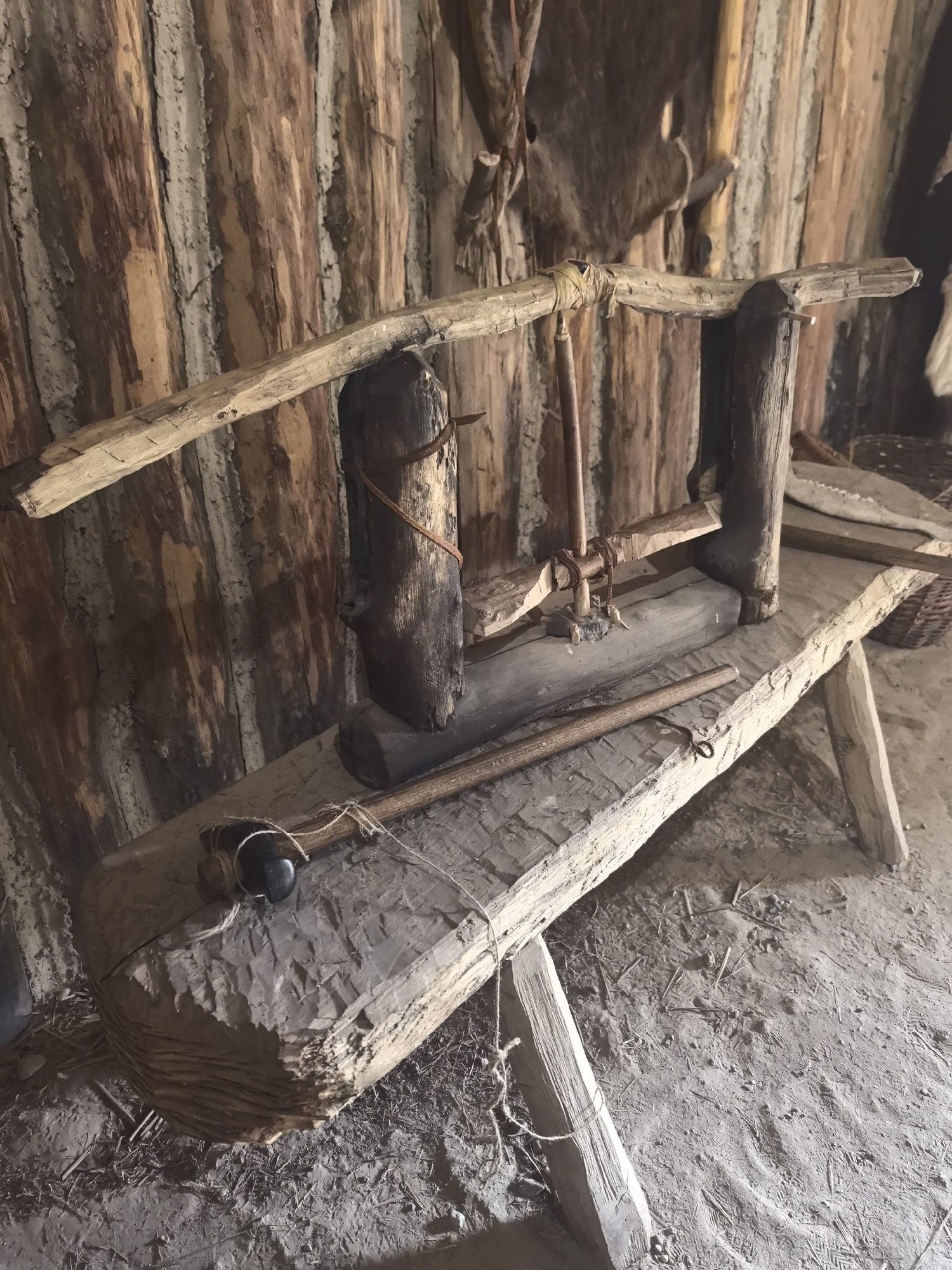 The image depicts an interior scene of what appears to be a historical dwelling, likely a reconstruction or a preserved structure. The walls are constructed from vertical wooden logs, with spaces between them filled with a lighter-colored material, possibly daub or clay. A rough-hewn wooden bench or platform is the central element of the foreground. On this bench, there is a primitive wooden apparatus, possibly for processing materials or as a tool. This apparatus consists of several wooden components, including a beam supported by uprights, a spindle-like element, and a lever. A long, slender wooden stick or rod lies across the bench in front of the main apparatus, with a loop of twine attached to one end, possibly for securing it. The floor is covered in dirt and debris, suggesting a rustic and aged environment. In the background, other wooden elements and possibly animal hides are visible, contributing to the primitive aesthetic. The lighting is natural and somewhat dim, typical of an interior without modern illumination. No people are present in the scene. The image does not provide any information about its location in the city of Камянюцкі сельскі Савет, Belarus.