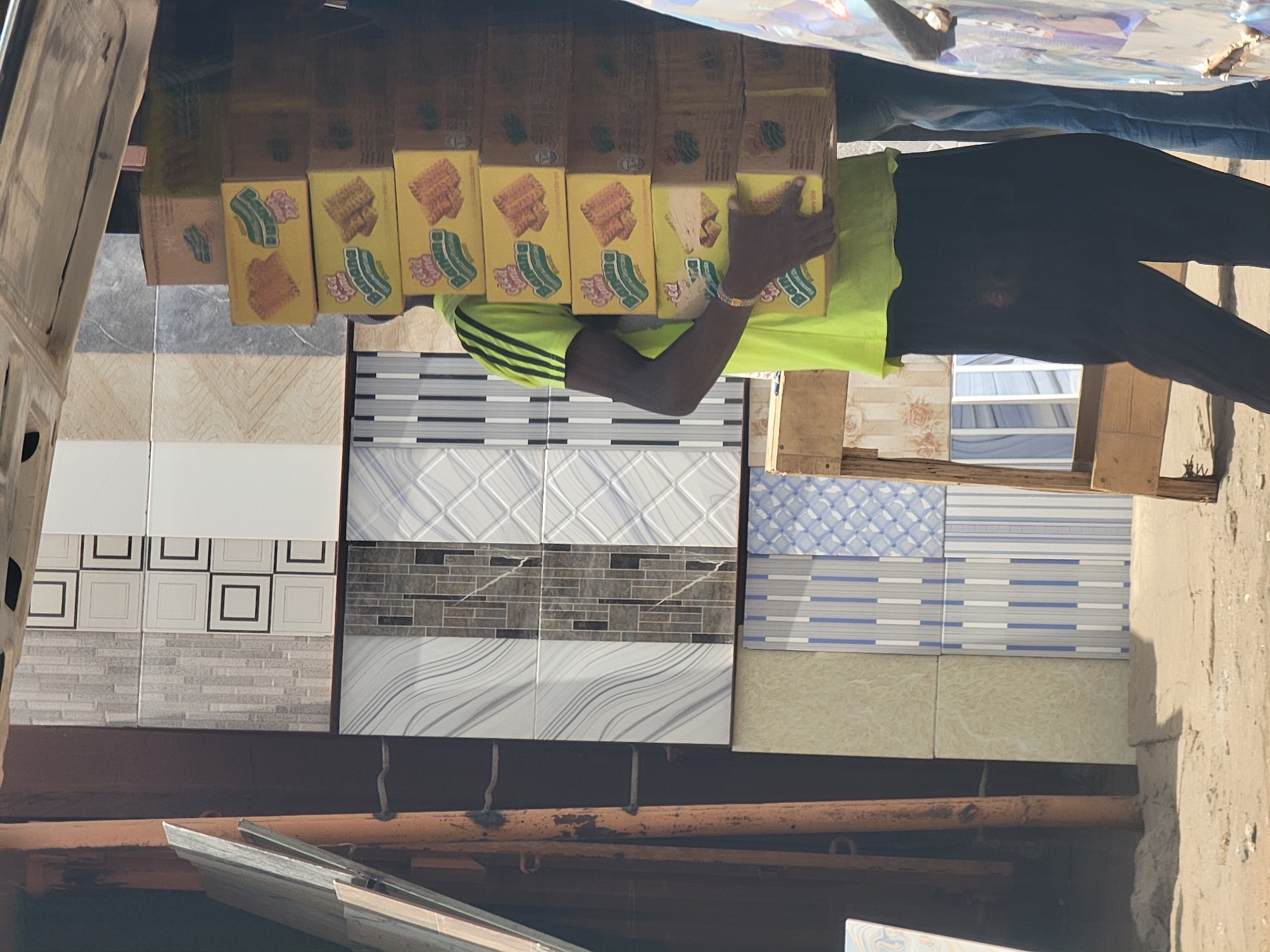 In Maiburiji, Nigeria, a man wearing a green t-shirt and black pants is holding a stack of yellow biscuit boxes. The boxes are stacked in front of a display of various ceramic tiles. The tiles are in different colors and patterns and are hung up for display. The scene is situated under a makeshift shelter or canopy. The environment appears to be a small business establishment or market stall.