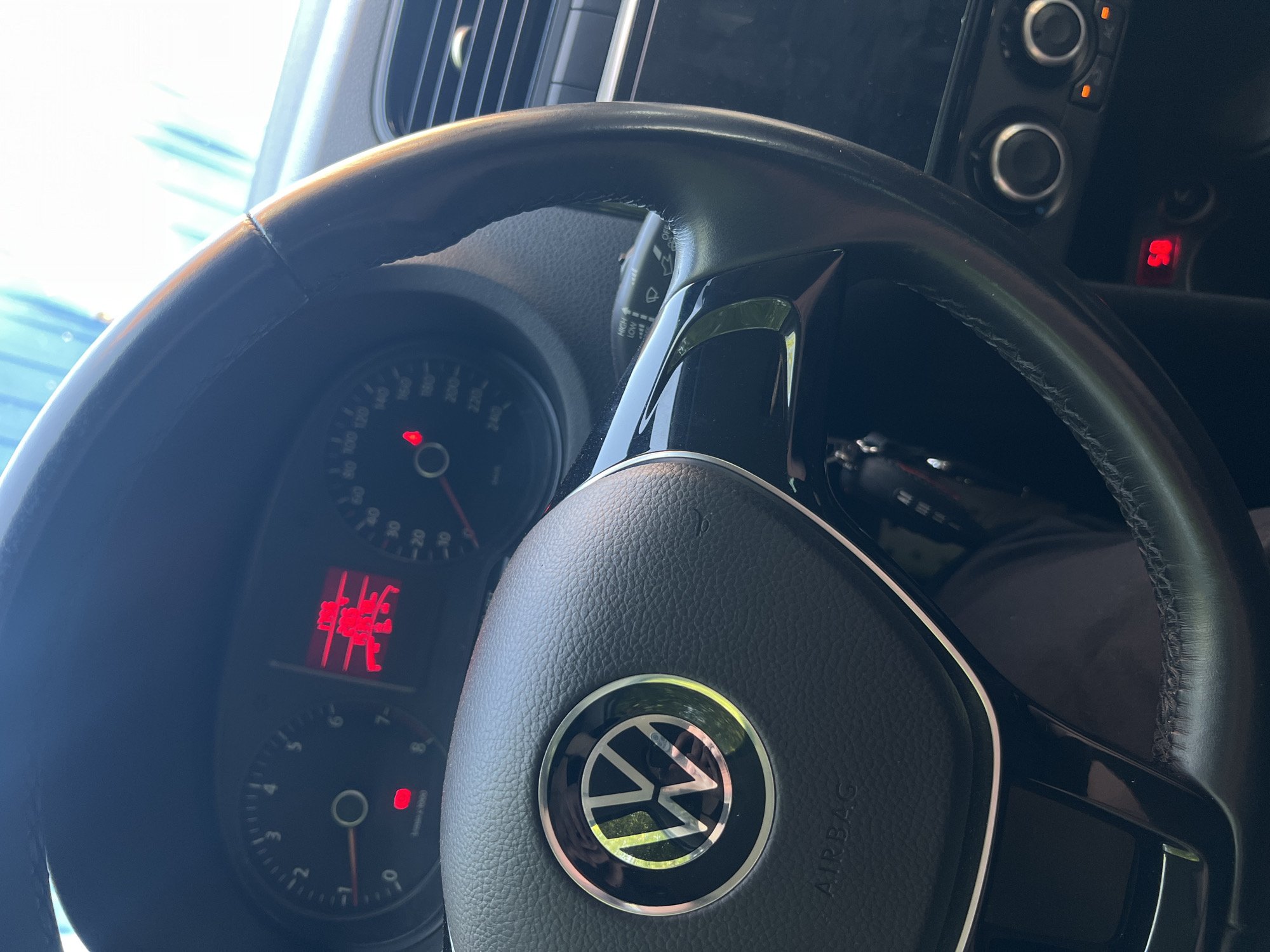 The image shows the interior of a Volkswagen car. The focus is on the steering wheel, displaying the VW logo and the "AIRBAG" label. The dashboard behind the steering wheel includes a speedometer and other gauges with illuminated red indicators and digital display. To the right of the steering wheel is a portion of the center console with various buttons and knobs. The location is unspecified.