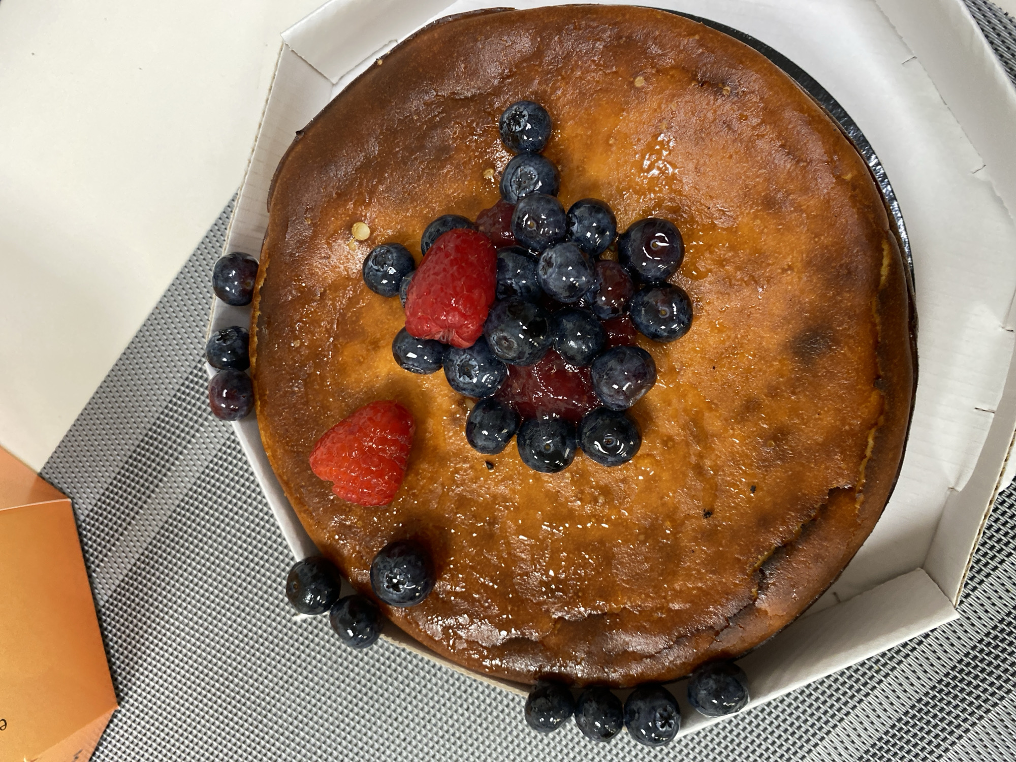 A cheesecake rests in a white cardboard box, adorned with blueberries and two raspberries arranged centrally on top. A reddish glaze or jam is visible amongst the blueberries. Some blueberries are scattered around the cake both inside and outside of the box. The cake appears to have a dark, baked crust. The box is placed on a surface with a gray and white checkered pattern. An orange-colored object is seen to the left of the cake box. The exact location in Saint Petersburg, Russia is not discernible from the image.