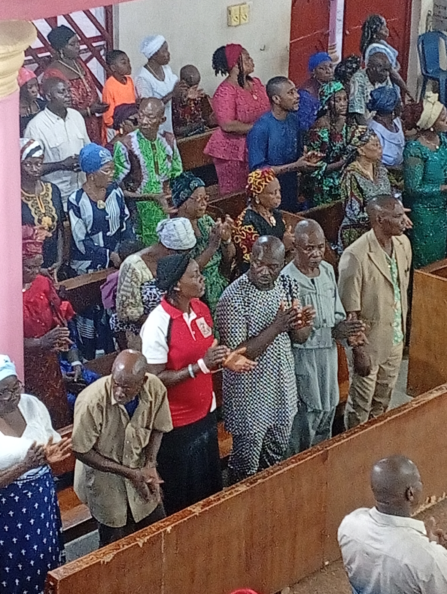 The image shows a crowded interior, possibly a church, filled with people of African descent. They are standing and clapping. Many are dressed in colorful and patterned clothing. Wooden pews are visible in the foreground. The location is in the city of Unknown, Unknown.