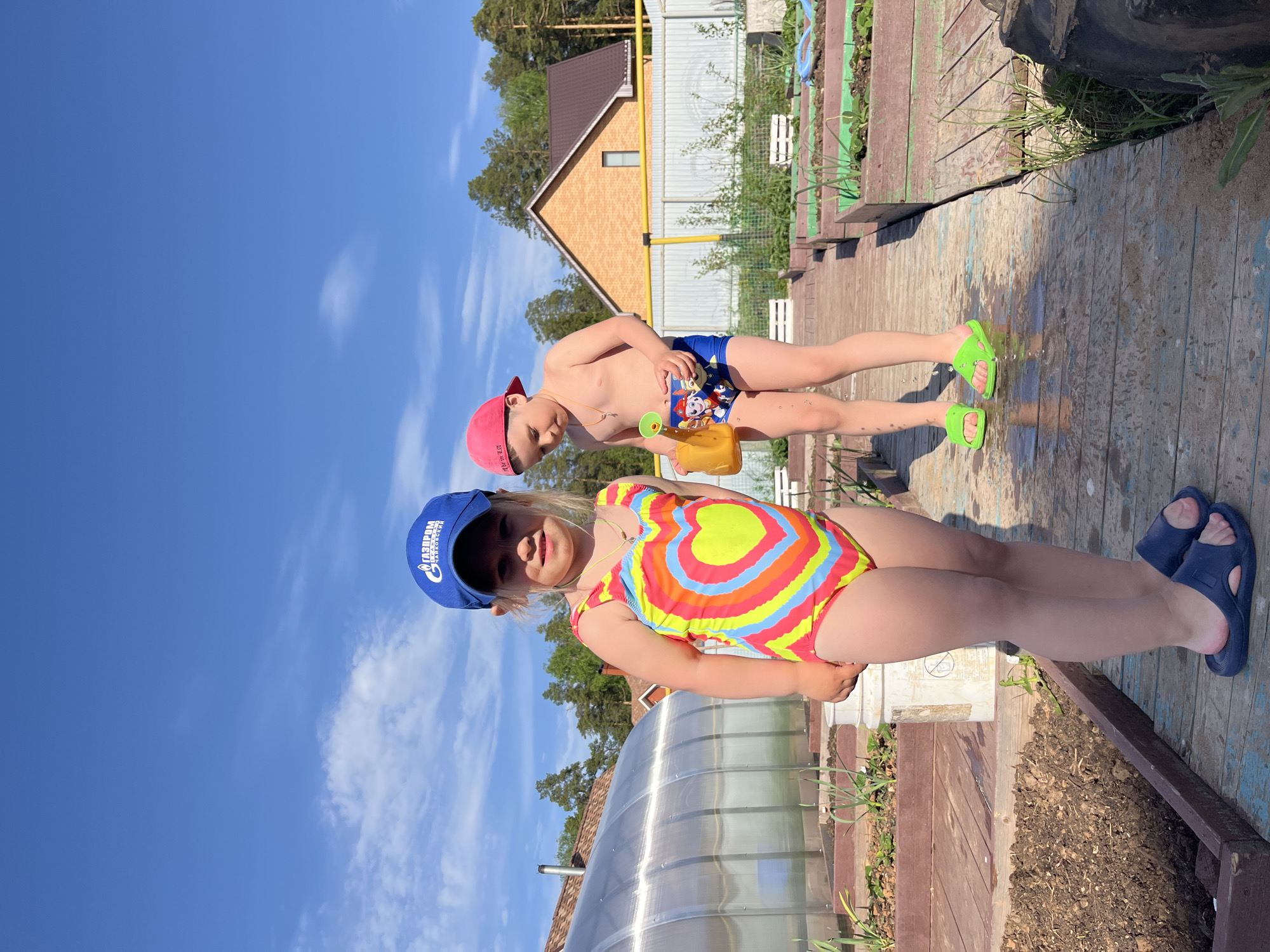 Two children stand outdoors on a sunny day, likely in a garden or backyard setting near the city of Chaikovsky, Russia. The foreground features a young girl wearing a blue cap with "Газпром Чайковский" (Gazprom Chaikovsky) printed on it, a colorful heart-patterned swimsuit, and blue sandals. Behind her stands a young boy, shirtless, wearing blue swim trunks and a red cap, holding a yellow toy watering can. A greenhouse is to the side of the girl.
The setting includes a wooden deck or walkway. A brick house with a brown roof is visible in the background. The sky is blue with scattered white clouds. The ground appears to be a mix of wooden planks, sand, and soil.