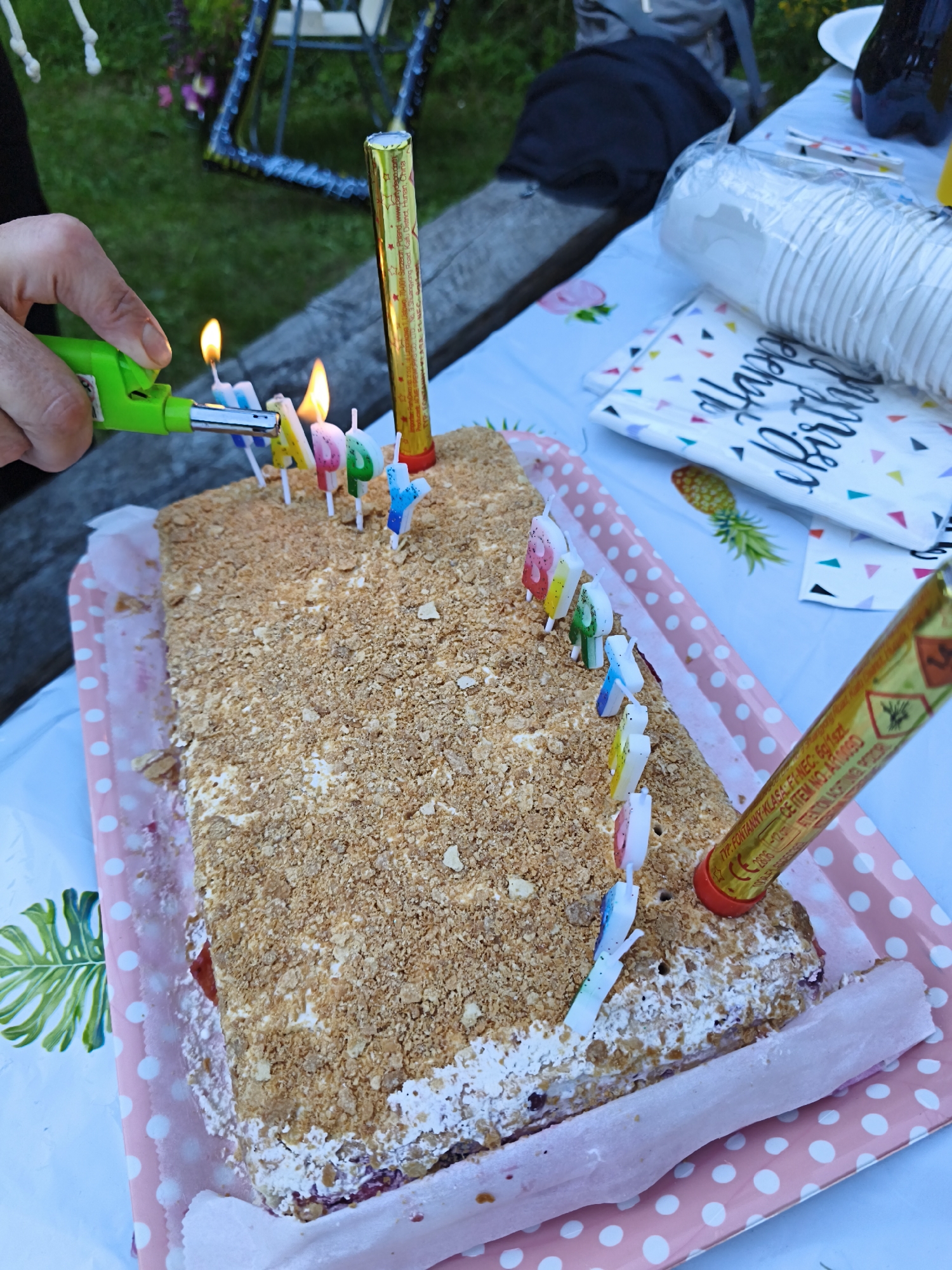 A rectangular cake with crumbled cookie topping and white frosting is on a pink and white polka dot tray. There are multiple birthday candles on the cake, some lit. A green lighter is being held by a hand to light one of the candles. To the right of the cake, there is a gold and red firework. The cake appears to be on a table covered with a blue and white patterned tablecloth, with other party supplies like a stack of white paper cups and a "Happy Birthday" napkin visible. The background shows greenery and a metal structure, suggesting an outdoor setting, possibly a garden. The overall scene depicts a birthday celebration.