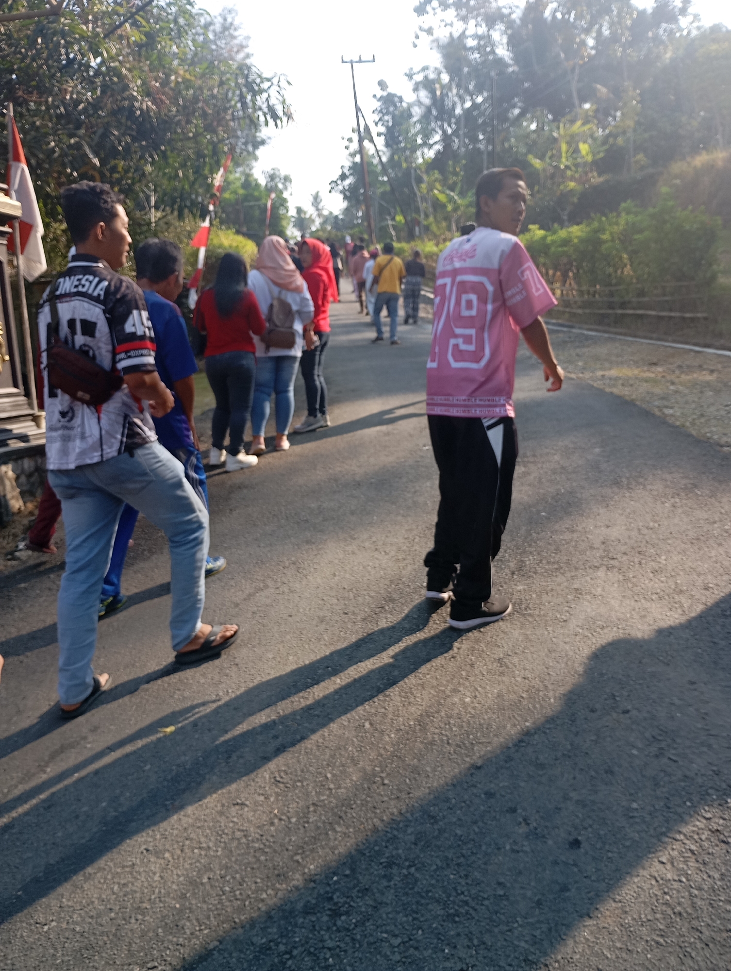 In Ketepung, Indonesia, a group of people are walking down a paved road lined with trees and vegetation. The sun is casting long shadows. On the left, a young man with short dark hair wears a black and white long-sleeved shirt with "ONESIA" written on it, and light blue jeans. He carries a dark brown bag across his chest. Next to him, a person in a blue shirt and jeans walks. Further down the road, a young man in a pink t-shirt with the number "79" on it and black pants is walking away from the camera. Several other individuals, some wearing headscarves, are also visible in the distance, appearing to be part of a procession or casual gathering. Two Indonesian flags are also visible on the left side of the image. The scene suggests a daytime outdoor activity in a rural or semi-rural setting.