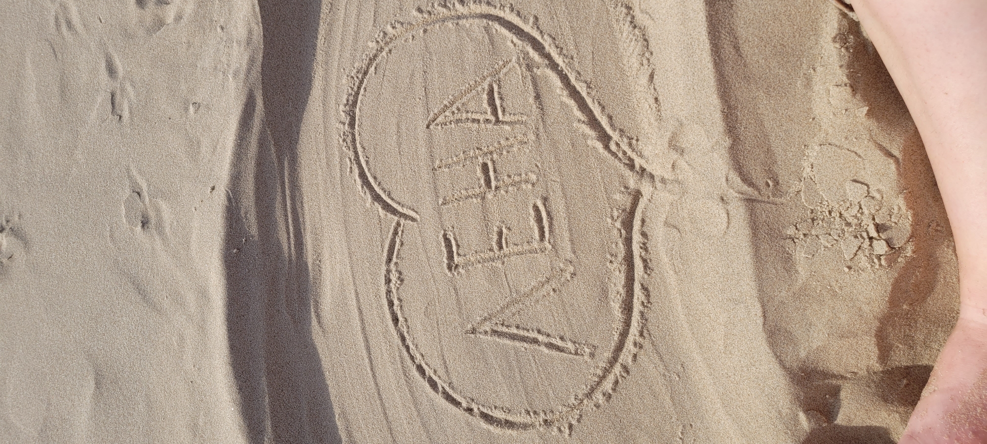 The image shows a heart drawn in the sand with the name "NEHA" written inside. The sand appears to be from a beach, and the lighting suggests it is daytime. There are also some indistinct shadows and a hint of human legs in the lower part of the image. The location is identified as the city of Riga, Latvia. This implies the drawing was made on one of Riga's beaches, such as the one along the Daugava River or on the coast of the Baltic Sea.