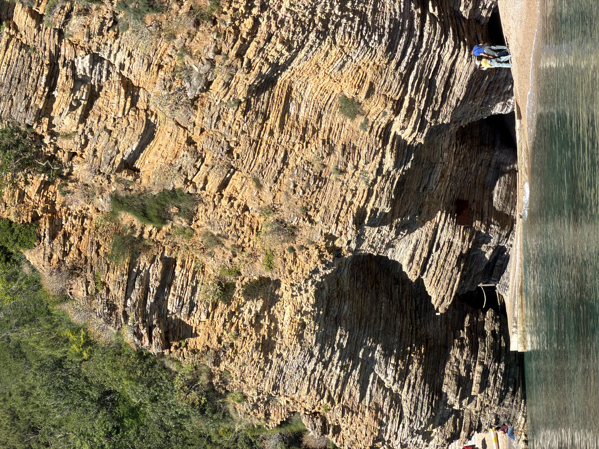 A cliff face on the coastline of Budva, Montenegro, is observed from a slightly rotated perspective. The cliff face is layered with sedimentary rock formations of varying sizes and shades of brown and tan. Vegetation consisting of green plants and shrubs is seen at the base and along the cliff's surface. Two people are positioned on a ledge along the cliff face, one wearing a blue shirt and the other wearing a yellow shirt. Water, likely from the sea, is visible alongside the cliff.