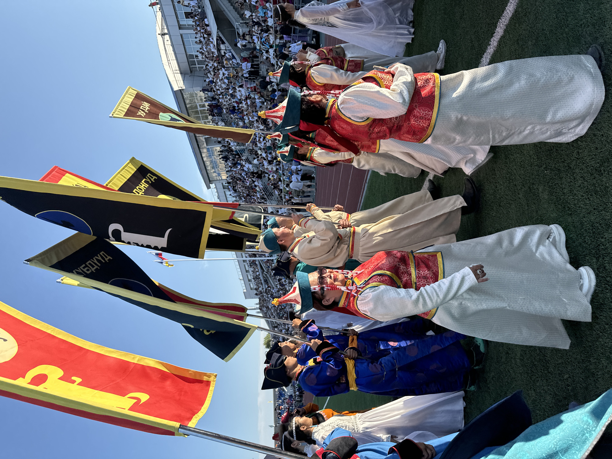 This media file captures a lively scene with people dressed in traditional attire, possibly participating in a cultural event or performance. The subjects are a group of individuals, including some who appear to be younger and others older, adorned in colorful, ornate clothing. The attire includes long, flowing skirts, decorated vests with intricate patterns in red and gold, and distinctive hats. Some individuals are holding or carrying flags, which are triangular and feature text in a non-Latin script, along with symbols.
The setting is an outdoor stadium with a green turf field, likely a sports ground. In the background, spectators fill the stands of the stadium, indicating a public gathering. The sky is clear and blue, suggesting a sunny day, and the lighting indicates it is daytime, likely late morning or afternoon. The overall atmosphere seems festive and celebratory. The event taking place could be a parade, a cultural festival, or a performance. The image is oriented vertically, with the subjects mostly seen from the waist down or in profile. The focus is on the costumes and the flags, with the background elements providing context to the scale of the event.