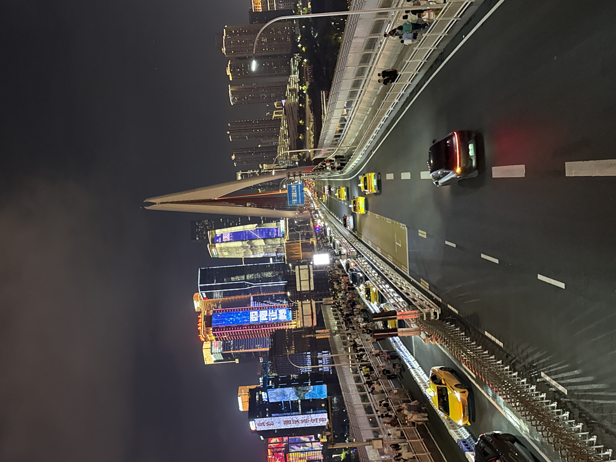 The image depicts a nighttime scene on a busy bridge in Yuzhong District, China, overlooking a cityscape illuminated by numerous bright skyscrapers. The bridge itself is wide, with multiple lanes of traffic and a pedestrian walkway. Several yellow taxis and other vehicles are visible on the road. A large crowd of people is walking along the pedestrian area, some observing the city skyline. The iconic architecture of a modern bridge with a prominent white pylon is featured, contributing to the dramatic urban landscape. The sky is dark and cloudy, with the lights from the buildings creating a vibrant contrast.