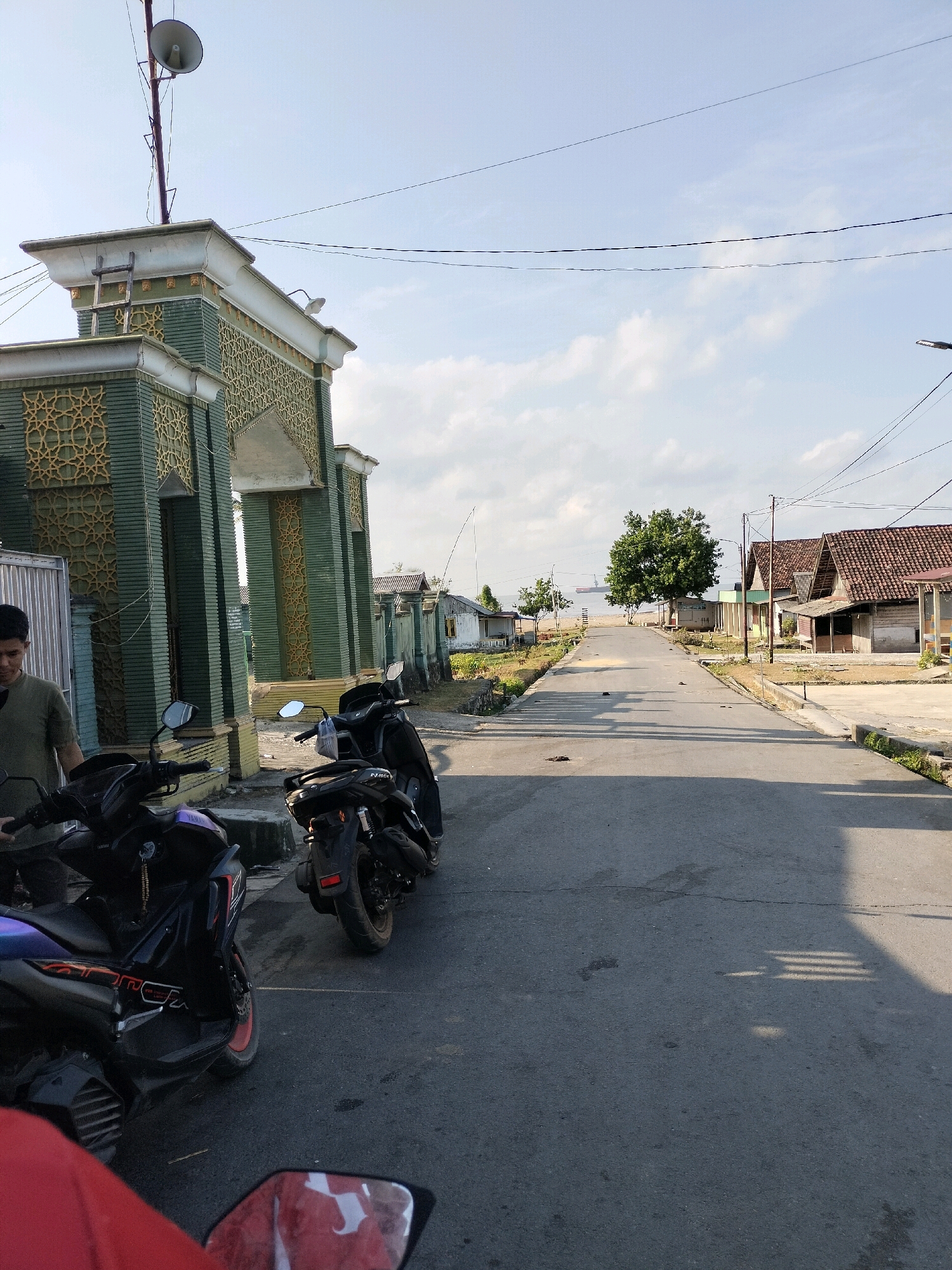 The image depicts a street scene in Bunati, Indonesia. On the left, an ornate green and gold gate stands with a loudspeaker mounted on a pole above it. Two motorcycles are parked in front of the gate. A man in a green shirt is standing to the left of the motorcycles. The street stretches forward, lined with simple buildings on the right, some with tiled roofs. In the distance, trees are visible, and further beyond, the sea with a ship. The sky is partly cloudy. The scene appears to be a quiet residential or coastal area.