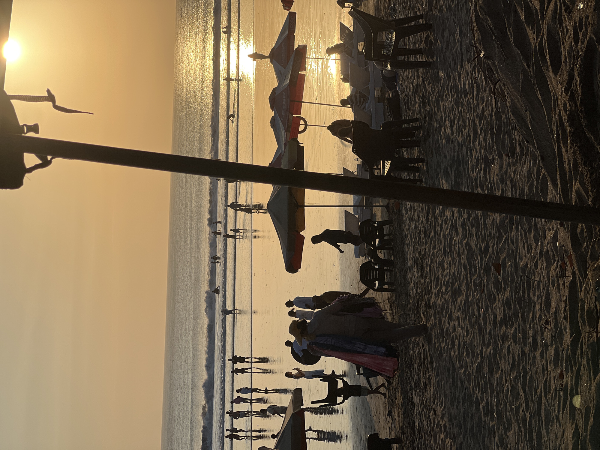 This image depicts a crowded beach scene in Kuta, Bali, Indonesia, during sunset. The sun is low in the sky, casting a warm, golden glow over the ocean and the sand. People are scattered across the beach, some wading in the shallow water, while others are walking or sitting on beach chairs under umbrellas. In the foreground, a person is walking away from the camera, carrying colorful fabrics. Several beach umbrellas and chairs are set up on the sand, some occupied. The ocean has gentle waves, and the wet sand reflects the colors of the sunset. The overall atmosphere is relaxed and lively, with many people enjoying the evening on the beach.