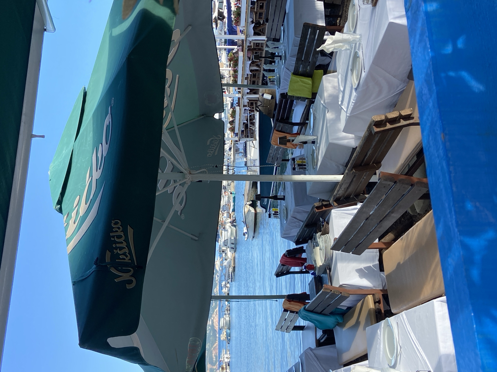 The image captures a vibrant outdoor dining area set up on a terrace overlooking a body of water, likely a harbor or bay in Budva, Montenegro, given the context. The scene is brightly lit under a clear blue sky, suggesting a sunny day.
In the foreground, numerous restaurant tables are neatly arranged, each covered with a crisp white tablecloth and set with plates and glasses. The seating consists of rustic wooden benches and chairs, some adorned with colorful cushions in shades of lime green and teal or light blue, adding splashes of color to the otherwise white and wood palette. Large green parasols are open, providing shade over the tables. One prominent parasol features the brand name "Nikšićko" in a decorative script, indicating a local beer brand.
Beyond the dining area, the tranquil blue water stretches out, dotted with several boats docked or anchored. In the distance, across the water, the coastline is visible with buildings and natural landscape, hinting at a bustling town or resort area. A solid blue structure, possibly part of the terrace railing or a wall, frames the right side of the image.
The overall impression is one of a peaceful, inviting restaurant awaiting customers, set against a picturesque coastal backdrop. No people are visibly present, suggesting either early morning preparations or a quiet moment before peak hours.