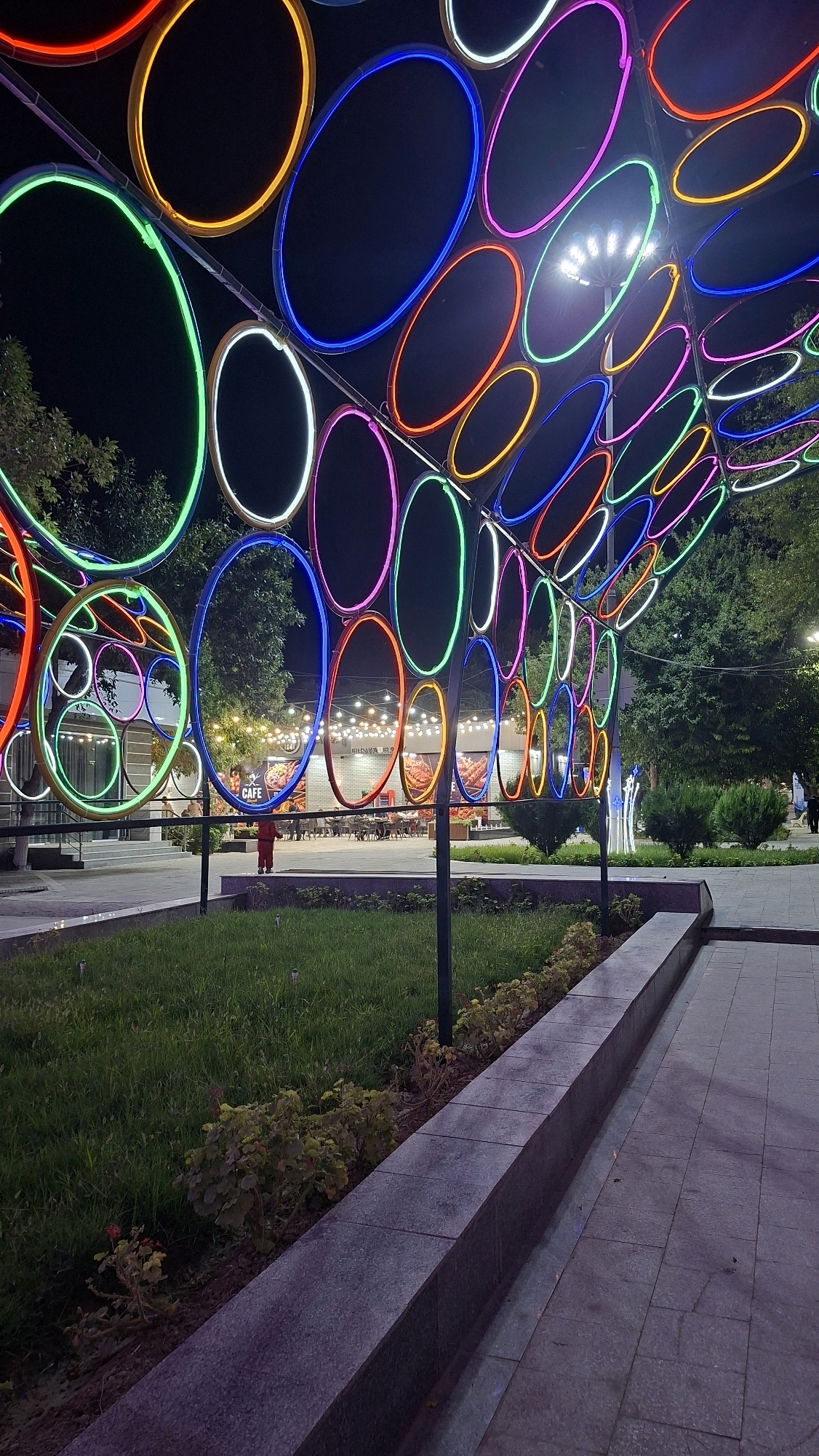 The image captures an outdoor scene at night, likely in a public space in Navoiy City, Uzbekistan, illuminated by vibrant, colorful neon ring lights forming an overhead canopy. These lights, in shades of red, orange, yellow, green, blue, and pink, create an abstract, futuristic atmosphere. Below the canopy, a patch of neatly mowed green grass is visible, bordered by a low, grey stone wall and a paved walkway. In the background, a row of buildings with illuminated windows suggests a commercial area, possibly a cafe or restaurant, with tables and chairs set up outside. A lamppost with multiple lights stands in the middle ground, further contributing to the illumination of the area. A single child, wearing a red outfit, stands in the center of the frame, facing away from the camera and towards the buildings. The overall impression is one of a lively, modern urban setting designed for evening entertainment or passage.