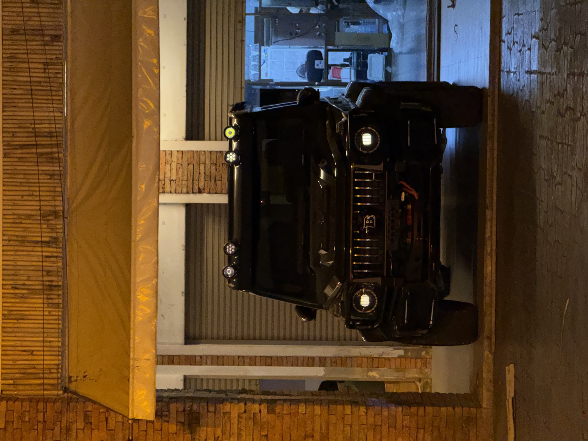 A black Mercedes-Benz G-Class SUV is parked in front of a building with a closed metal garage door. The SUV has a custom grille with a "B" logo and additional lights mounted on the roof. The building appears to be made of brick and has a yellow awning. The scene is lit by artificial lights, casting shadows on the ground, which appears to be paved. The overall impression is that of a dark, possibly nighttime scene in an urban environment, specifically Baan Bo Phut, Thailand, as indicated by the context.