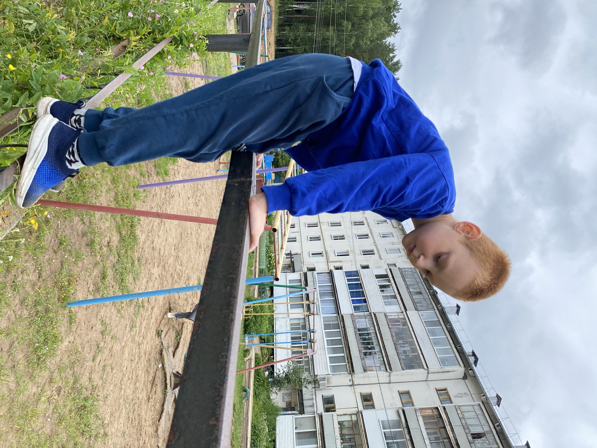 A young boy with ginger hair is balancing on a horizontal metal bar on a playground. He is wearing a blue sweatshirt and dark blue sweatpants, with blue and white sneakers. His gaze is directed downwards. Behind him, a multi-story apartment building with numerous balconies and windows is visible, characteristic of Soviet-era architecture. The playground area consists of sand and some greenery. The sky is overcast. The scene is situated in the city of Noviy, Russia.