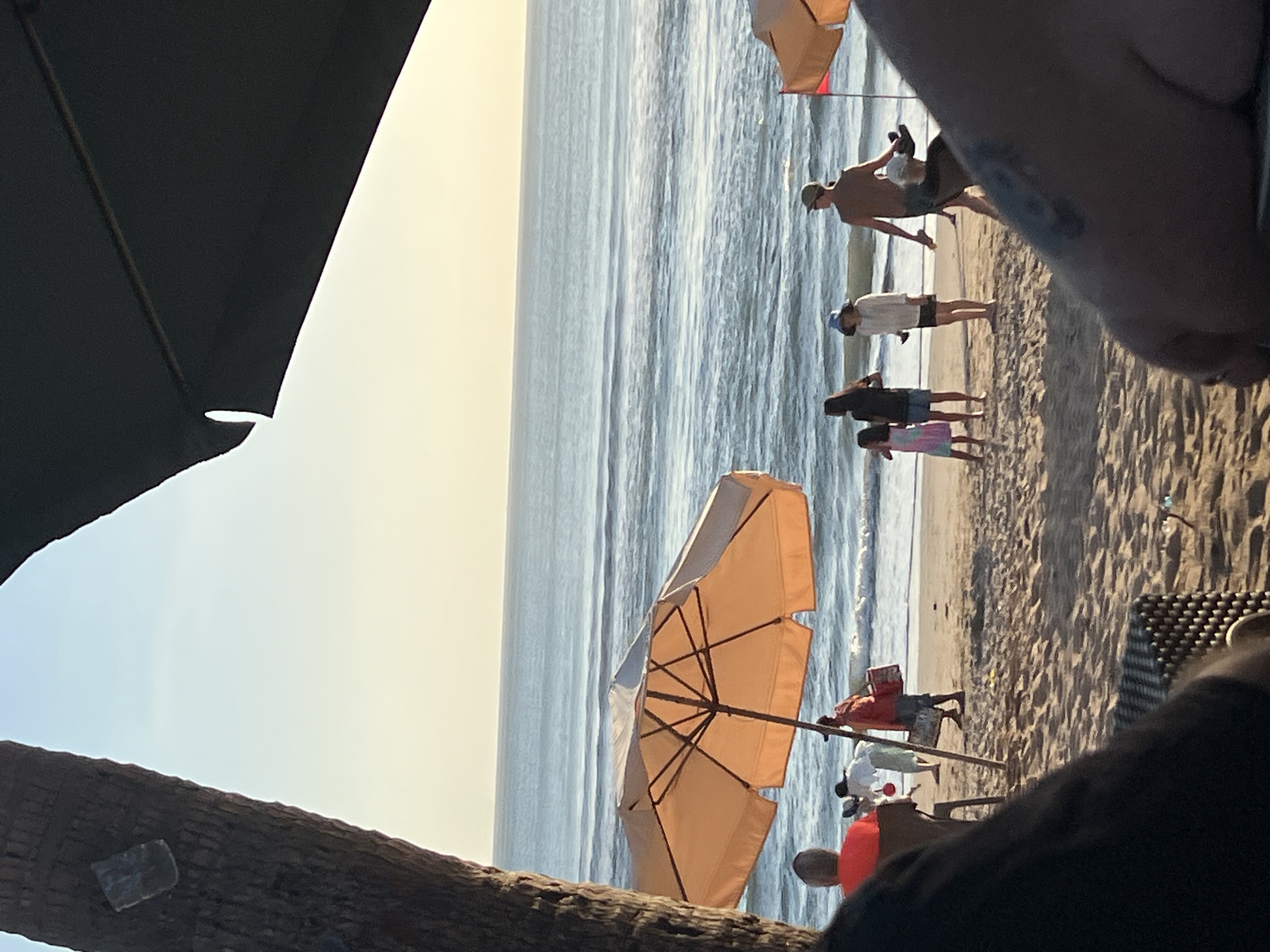 An eye-level, medium shot captures a beach scene in Kuta, Indonesia, under a clear, soft golden sky as the sun begins to set. In the foreground, the blurred out-of-focus view from a seating area looking towards the ocean dominates the bottom portion of the frame. The middle ground is a sandy beach where several people are present. A large orange umbrella is set up on the sand, with a portion of another umbrella visible in the upper right corner. Several individuals are depicted walking and interacting on the beach, some appearing to be vendors. The ocean stretches out to the horizon, with gentle waves rolling in. The overall atmosphere is serene and relaxed, indicative of a tranquil evening by the sea in Kuta.