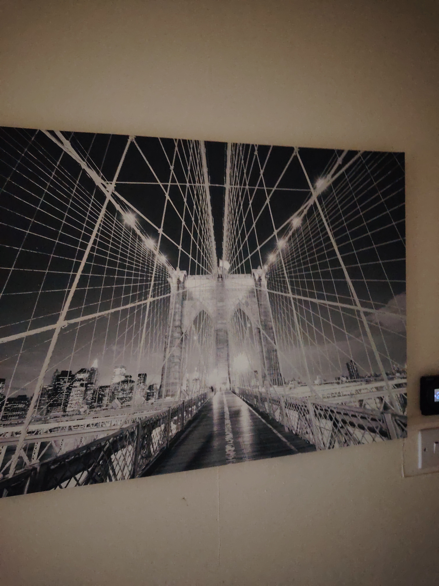 The image shows a large, black and white canvas print of the Brooklyn Bridge at night, mounted on a light-colored wall. The print is a wide landscape orientation. The bridge's suspension cables are prominently displayed, converging towards the center of the frame. The towers of the bridge are visible, illuminated by lights. In the background, a city skyline with numerous illuminated buildings is depicted. The foreground of the print shows the roadway of the bridge. The print is slightly angled on the wall, with the top edge appearing to be higher on the left than the right. To the right of the print, a white electrical socket and a small black device with a digital display are visible on the wall. The wall itself appears to have a textured surface. The lighting in the scene is dim, with the primary light source being the illumination from the city skyline and the bridge itself as depicted in the artwork. There are no people or discernible actions within the print. The location is identified as Huntingdonshire, United Kingdom, although the artwork itself depicts New York City.