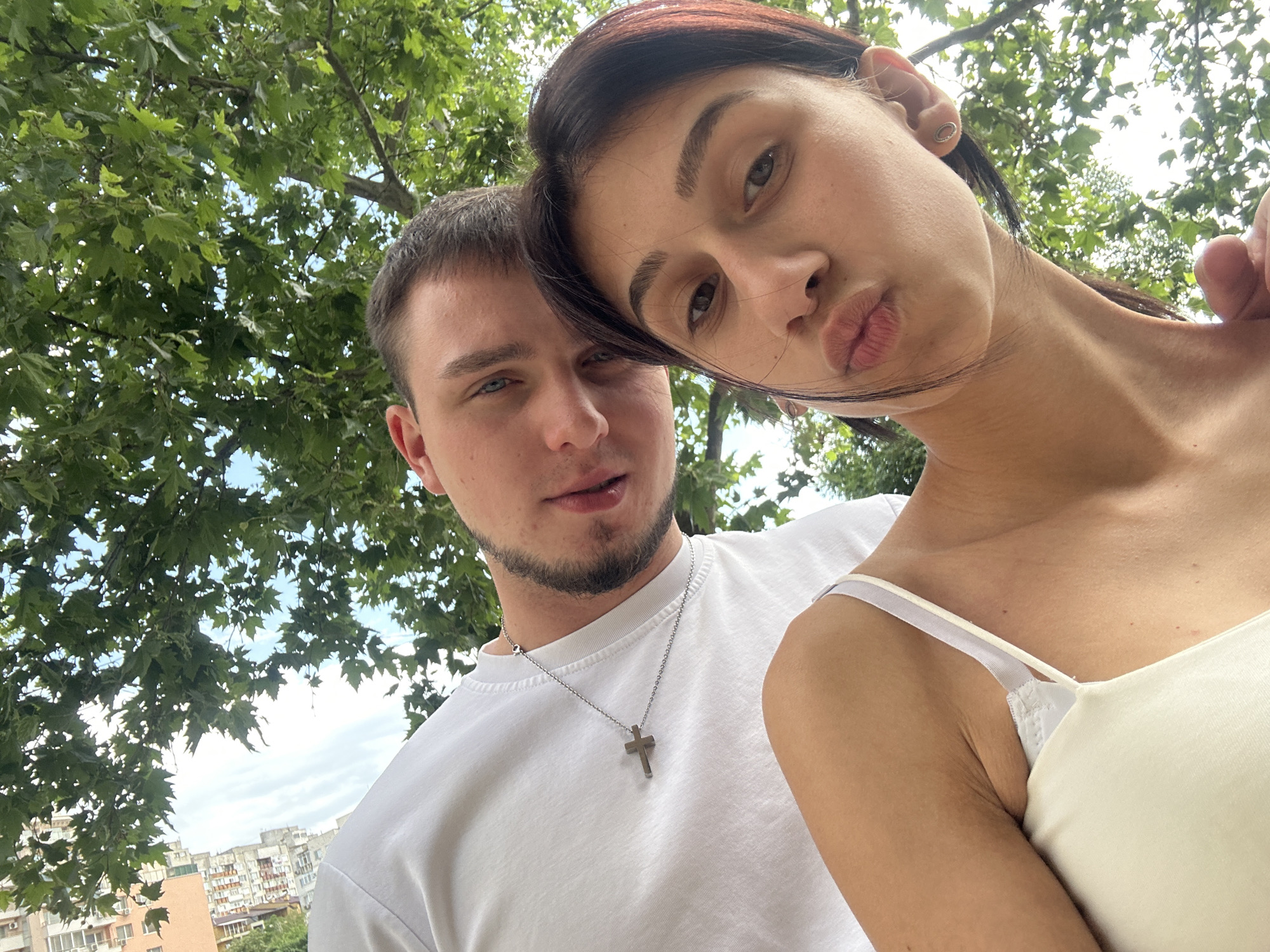 A selfie shows a man and a woman under a tree, possibly in Plovdiv, Bulgaria, given the presence of buildings typical of the region in the background. The woman, positioned closer to the camera, is making a pouting expression. She has dark hair and is wearing a light-colored tank top with thin straps. The man, behind and slightly to the left of her, has short hair and a beard and is wearing a white t-shirt with a silver cross necklace. The environment includes green leaves from the tree overhead, a glimpse of a cloudy sky, and the urban landscape of Plovdiv in the distance, with apartment buildings.