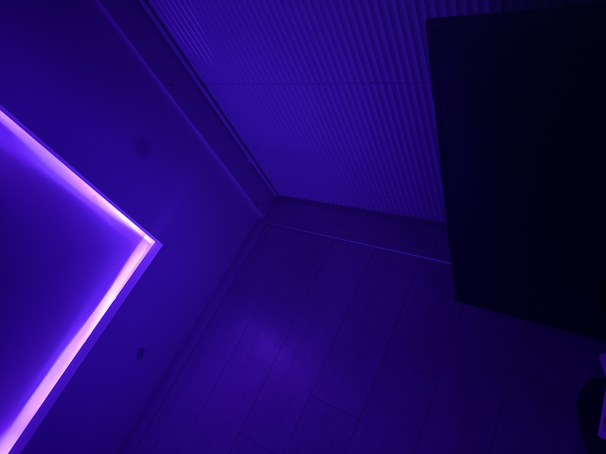 The image shows an indoor scene illuminated by purple light, with a modern ceiling design featuring integrated lighting. On the left, a wall appears to be paneled with wood. On the right, there are window blinds. In the foreground, a dark, flat object, possibly a piece of furniture or a screen, is visible. The lighting creates a dramatic and atmospheric effect, with strong shadows and highlights. The scene appears to be located in a contemporary interior space. There are no people or specific landmarks visible that would indicate the city of Burgas, Bulgaria.