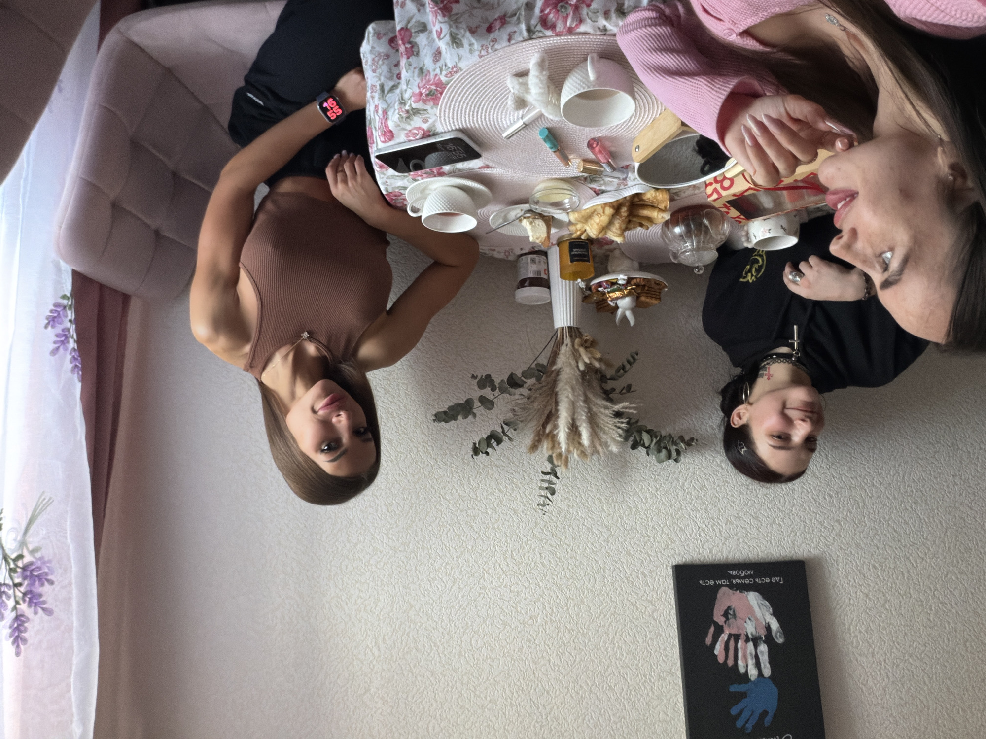 Two women are seated at a table in an indoor setting. A floral tablecloth covers the table, on which are an assortment of teacups, a teapot, pastries, and cosmetics. One woman wears a brown top and a smartwatch, while the other is in a pink sweater. The scene appears to be a tea party setting. In the background, there is a wall with a framed piece of art or a book with an image of handprints on it. Based on the style of decor and the items present, it's likely a domestic setting, possibly a home in Russia. It's difficult to pinpoint the specific location within Saint Petersburg without additional information, but it is most likely in a living area or dining room.