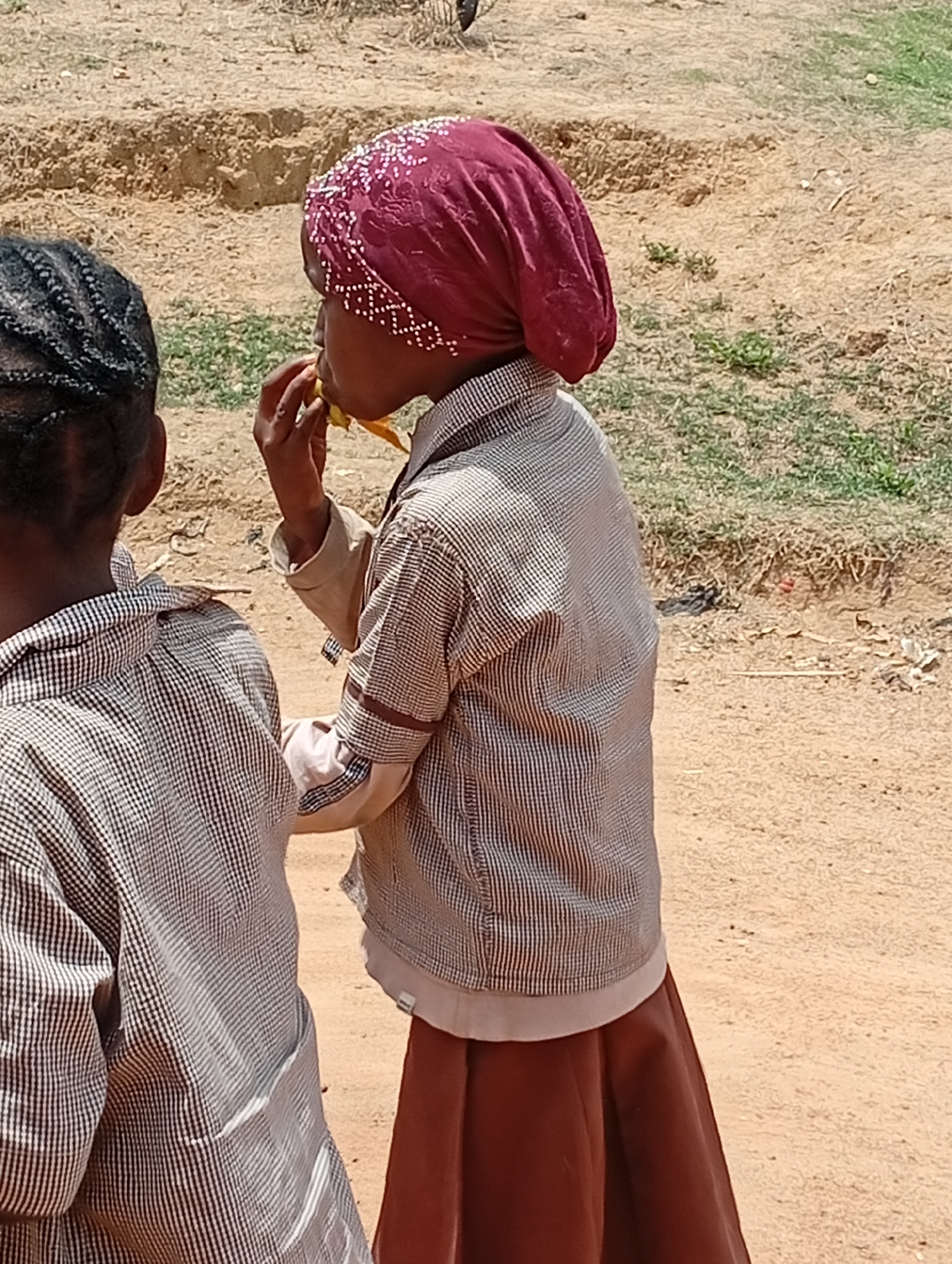In Barakin Akawo, Nigeria, two children are standing outdoors on a dirt ground. The child on the right is eating, with food in their hand and near their mouth. They wear a maroon head covering with small white embellishments, a plaid shirt with brown accents, and a brown skirt. The second child, partially visible on the left, has braided hair and wears the same plaid shirt as the first child. The background features a grassy area and dirt terrain.