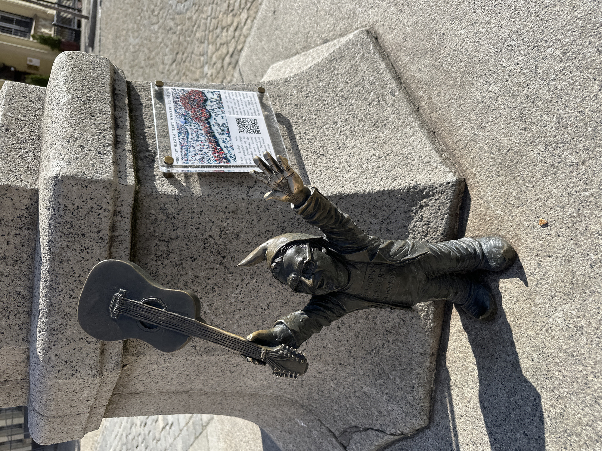 The image features a bronze statue of a gnome with a guitar emerging from a stone wall. Above the statue is a plaque with text in Polish that says "GITAROWY REKORD GUINNESSA," "WROCŁAW GITARY! WROCŁAW GUITAR GUINNESS RECORD FOR GUITAR," and other information. The statue is located in Wrocław, Poland, known for its gnome statues. The gnome has a pointed hat and is holding a guitar in its left hand, with the right arm extended.