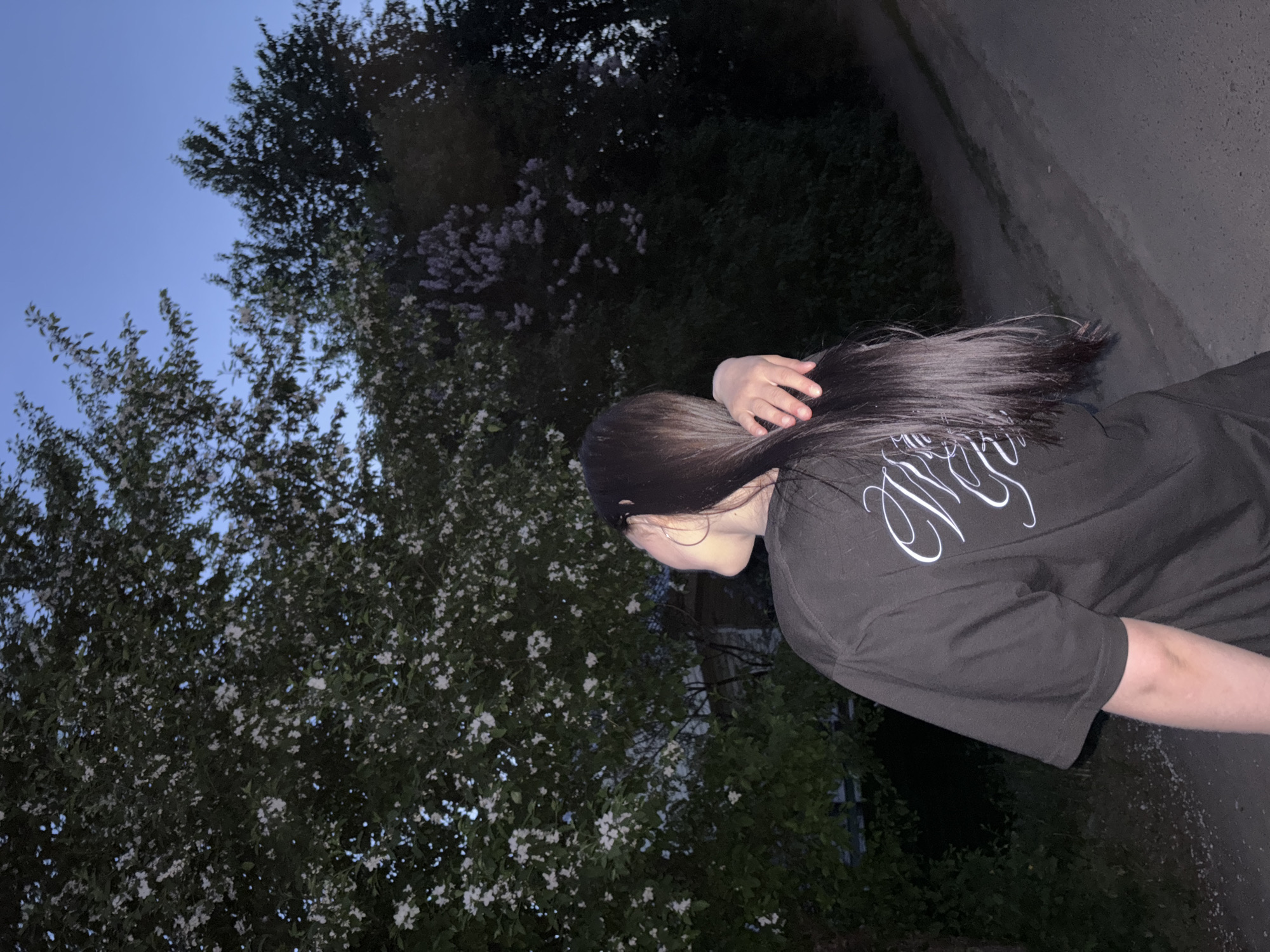 A woman with long, dark hair is standing next to a tree with white flowers, on what appears to be a paved road or sidewalk. The woman is wearing a dark-colored t-shirt with white lettering on the back. The background features a mix of trees and shrubs, and the sky is dusky blue, suggesting twilight. The scene is in Фоки, Russia.