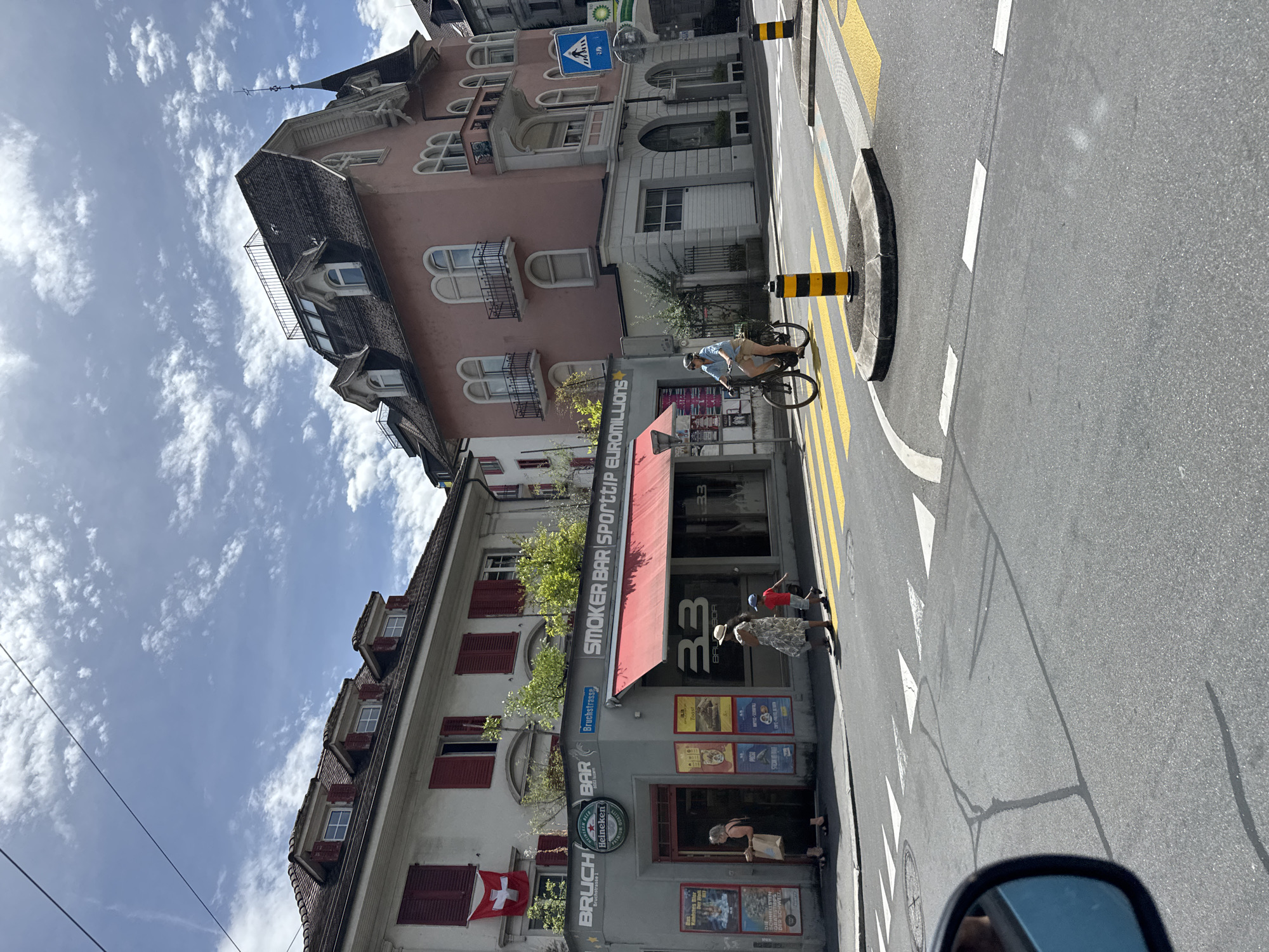 In Lucerne, Switzerland, on a sunny day with scattered clouds, a street scene unfolds at an intersection. A white building with red shutters and a Swiss flag hanging prominently on its facade stands on the left. Adjacent to it, a corner establishment displays signage for "BRUCH BAR HEINEKEN" and "SMOKER BAR SPORTTIP EUROMILLIONS" with a red awning. Posters adorn the exterior of the bar.
On the right side of the intersection, a distinct pink building with a complex roofline and multiple balconies dominates the background. In the foreground, a woman wearing a blue shirt and khaki shorts rides a bicycle across a pedestrian crossing marked with yellow and white stripes. Two other individuals, a woman and a child, are visible near the corner establishment, walking on the sidewalk. The street surface shows clear road markings, including a pedestrian crossing and lane dividers. The overall environment suggests a vibrant urban setting within Lucerne.