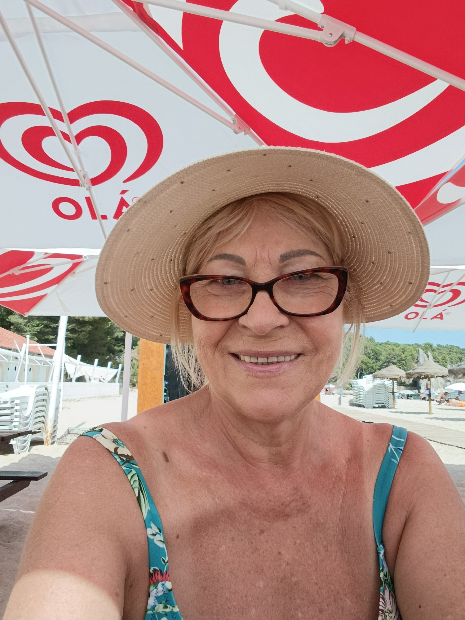 This is a selfie of a middle-aged woman wearing a straw hat and glasses, taken on a beach. She is smiling and looking at the camera. The background shows a beach with umbrellas, lounge chairs, and people enjoying the sun. The woman is wearing a floral swimsuit. The image appears to be taken in Setúbal, Portugal.