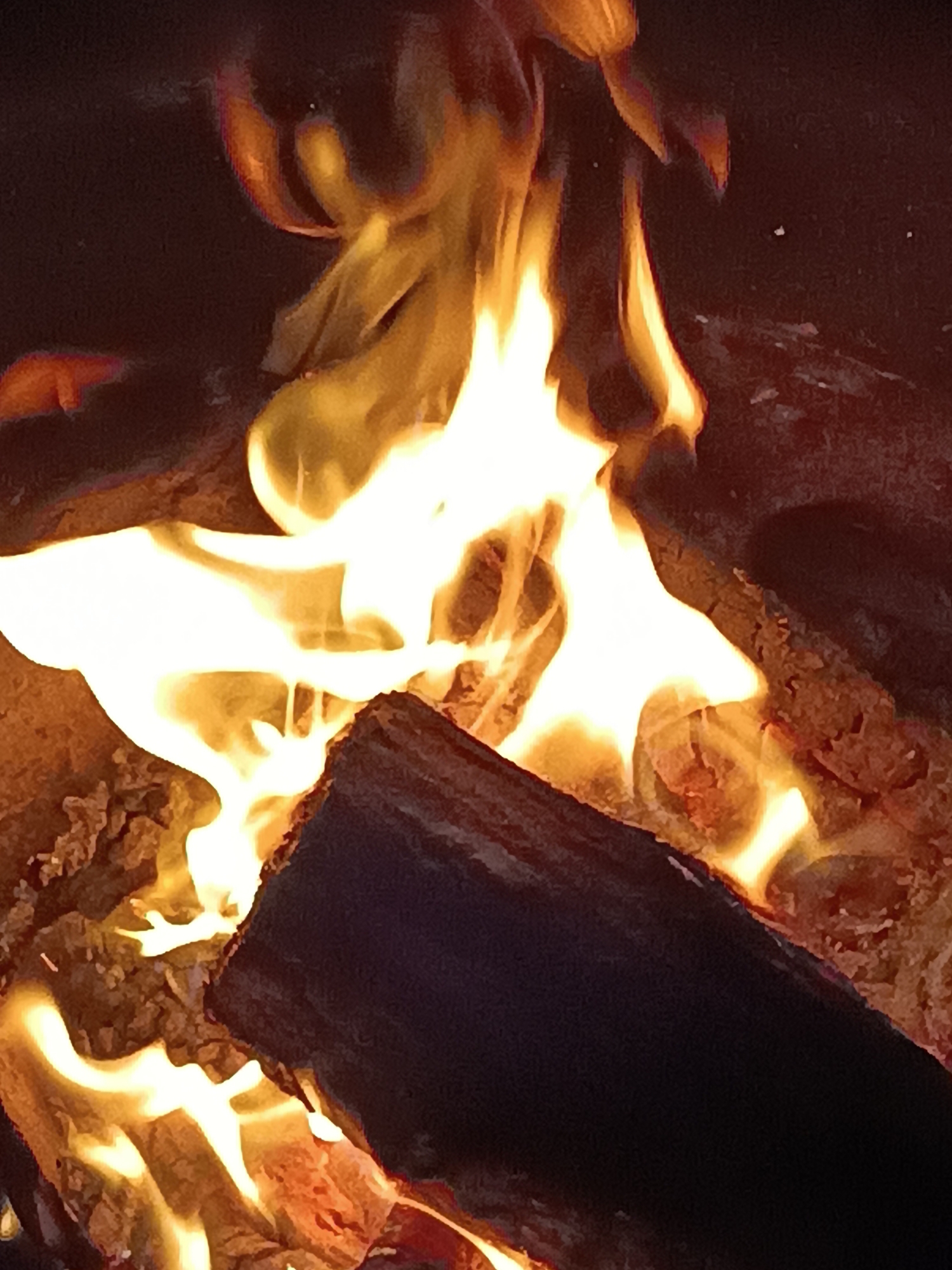 The image is a close-up shot of a fire burning in what appears to be a fireplace or fire pit. The flames are bright yellow and white, licking around dark, textured logs. The background is dark and out of focus, emphasizing the fiery activity. The scene suggests warmth and light. There is no visible indication of the city of Viadana, Italy, or any specific location. The image focuses solely on the fire and burning wood.