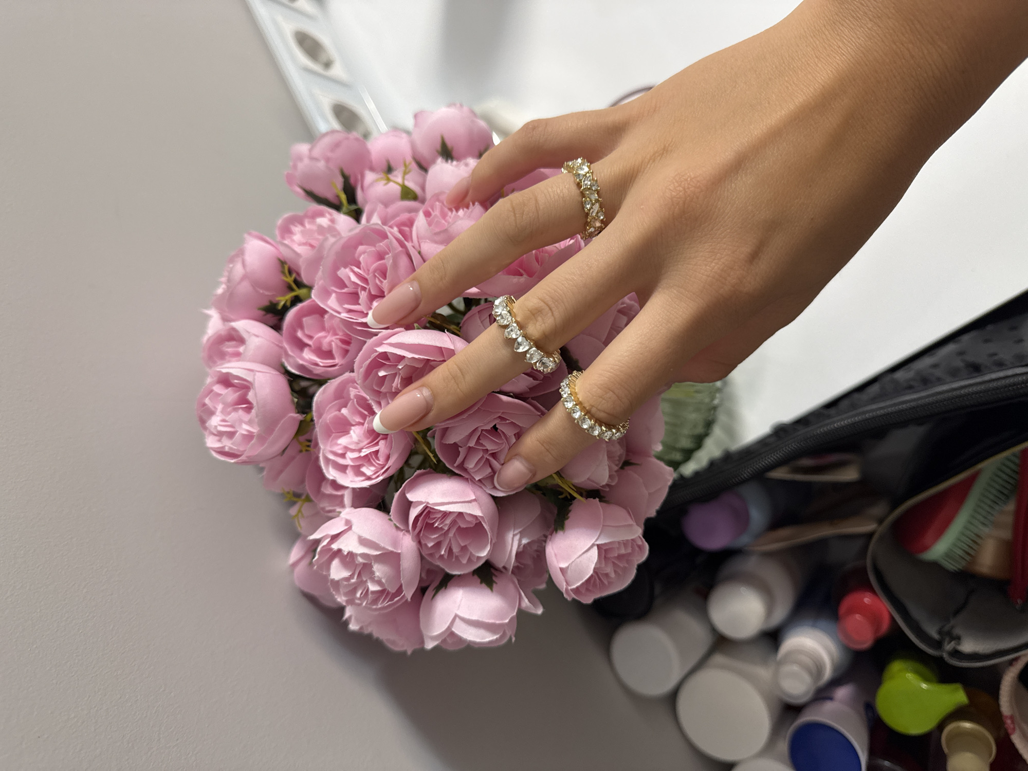 The image depicts a person's hand adorned with three gold and diamond rings, gently touching a bouquet of pink roses. The hand has manicured nails with a French tip design. To the right of the roses, there's a partially visible black makeup bag overflowing with various cosmetic bottles and containers. The background consists of a white surface on top and a grey wall on the left. There is no indication of a location in Burgas, Bulgaria, or any city. The focus is on the hand, rings, roses, and makeup bag, implying a beauty or personal care theme.