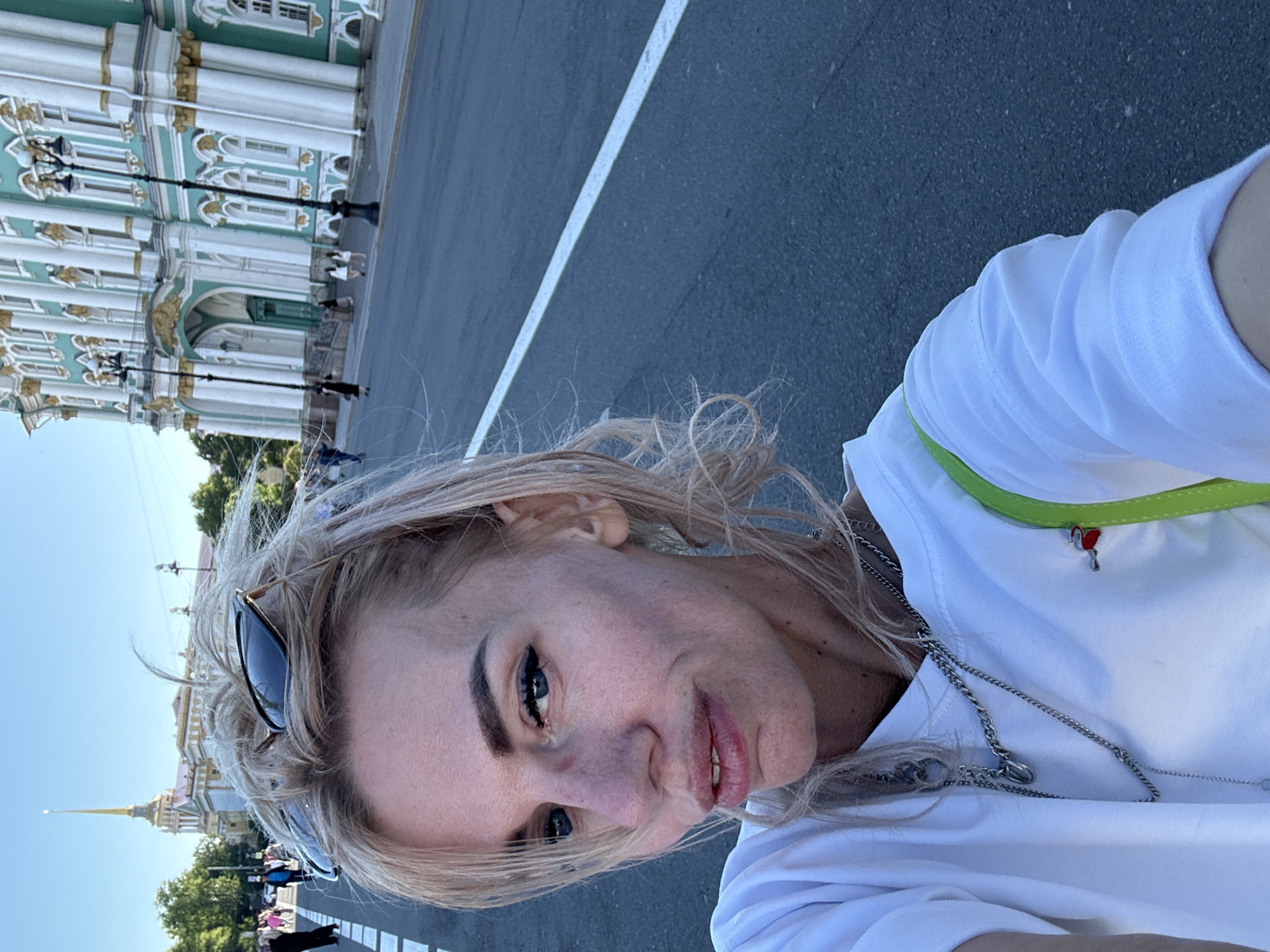 A young blonde woman is taking a selfie outdoors in the city of Saint Petersburg, Russia, in front of the Hermitage Museum. She is wearing a white t-shirt and multiple silver necklaces, and has sunglasses on top of her head. The Hermitage Museum, a former imperial palace, is easily recognizable by its distinctive green and white facade and classical architecture. The woman is standing on a paved road with a white line marking. In the background, other people are walking along the sidewalk. The sky is clear and blue, indicating a sunny day.